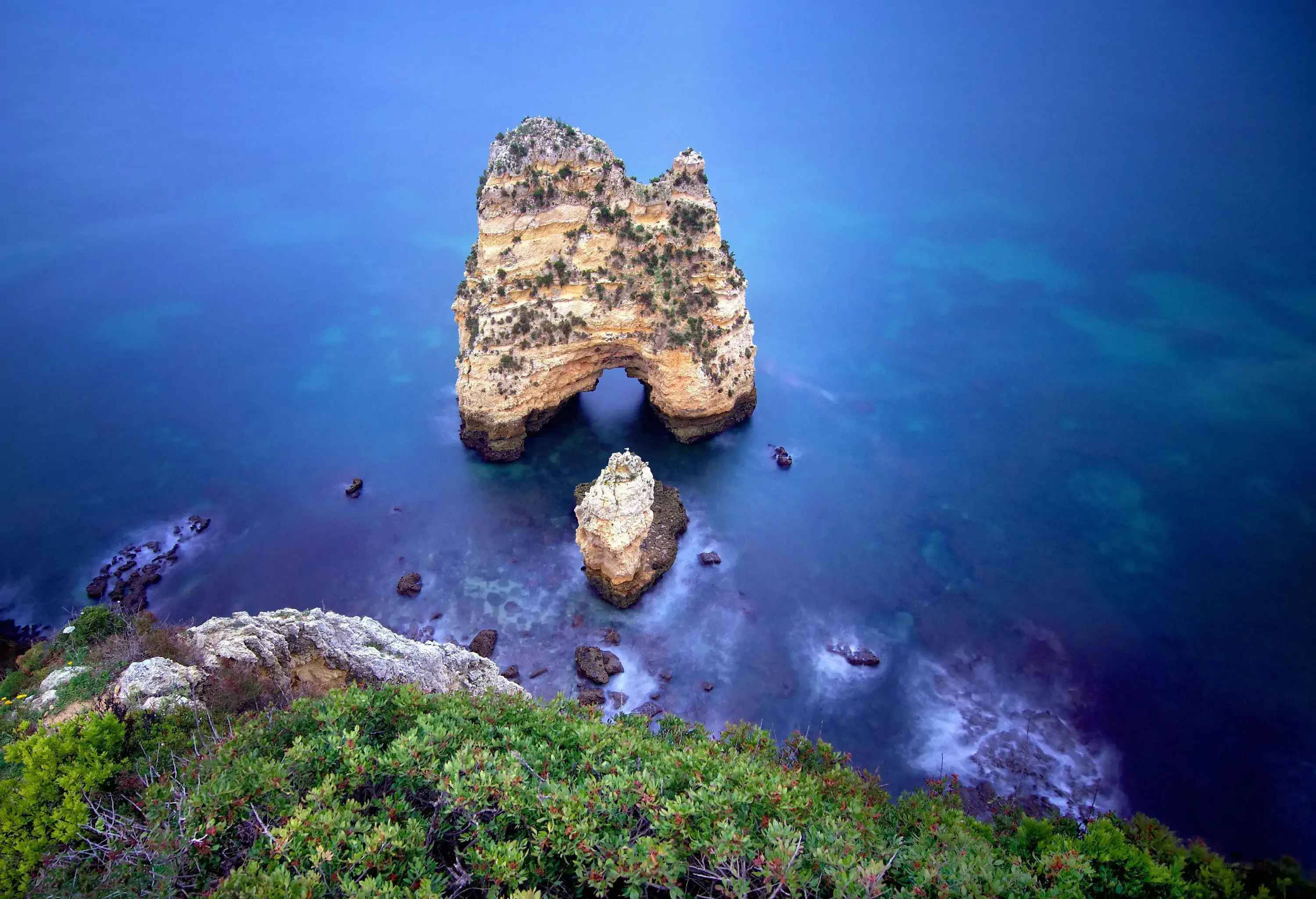 A natural rock arch in the middle of the blue sea next to a lush rocky island.