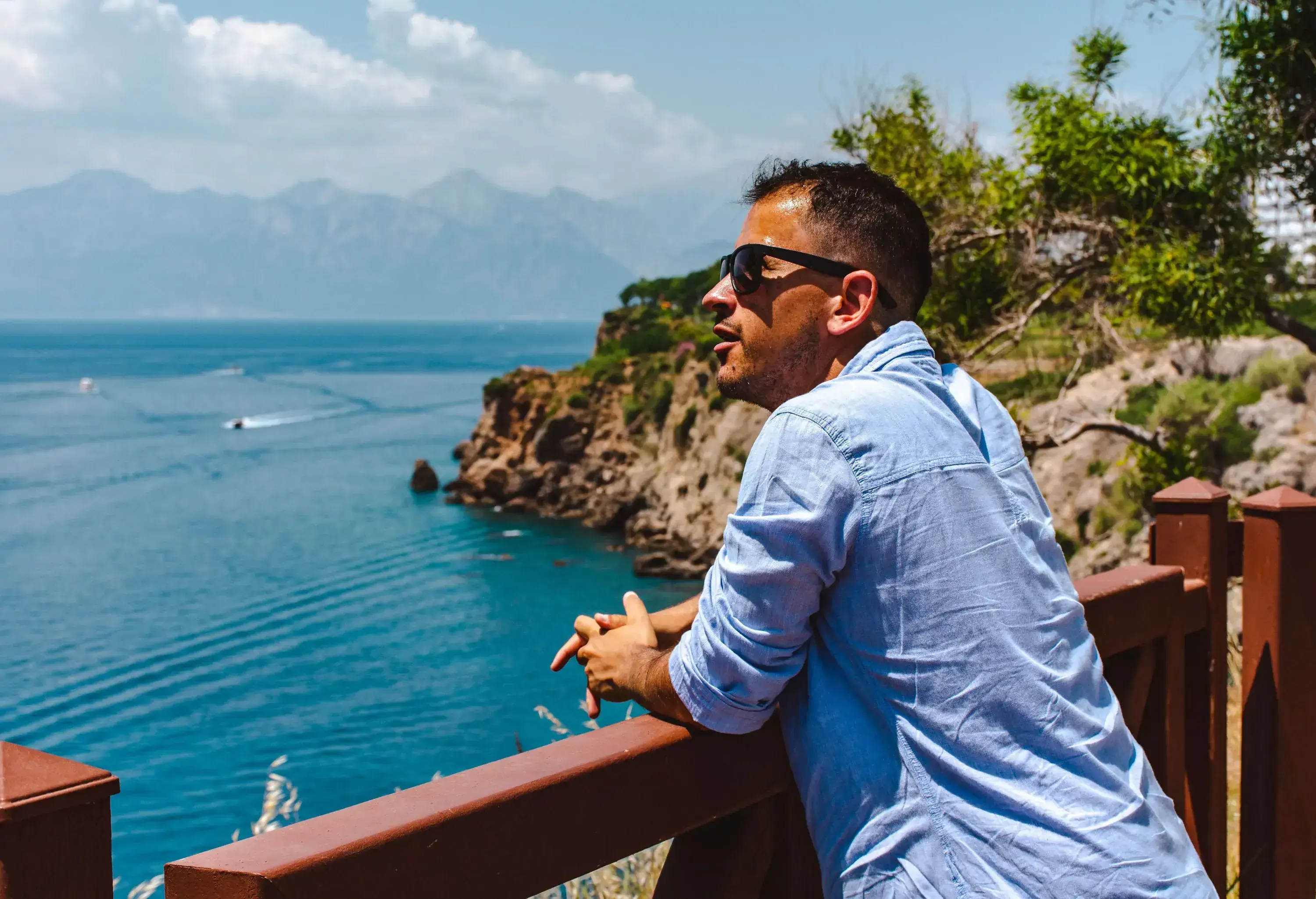 Person in sunglasses rests on a railing, enjoying a panoramic ocean and mountain view.