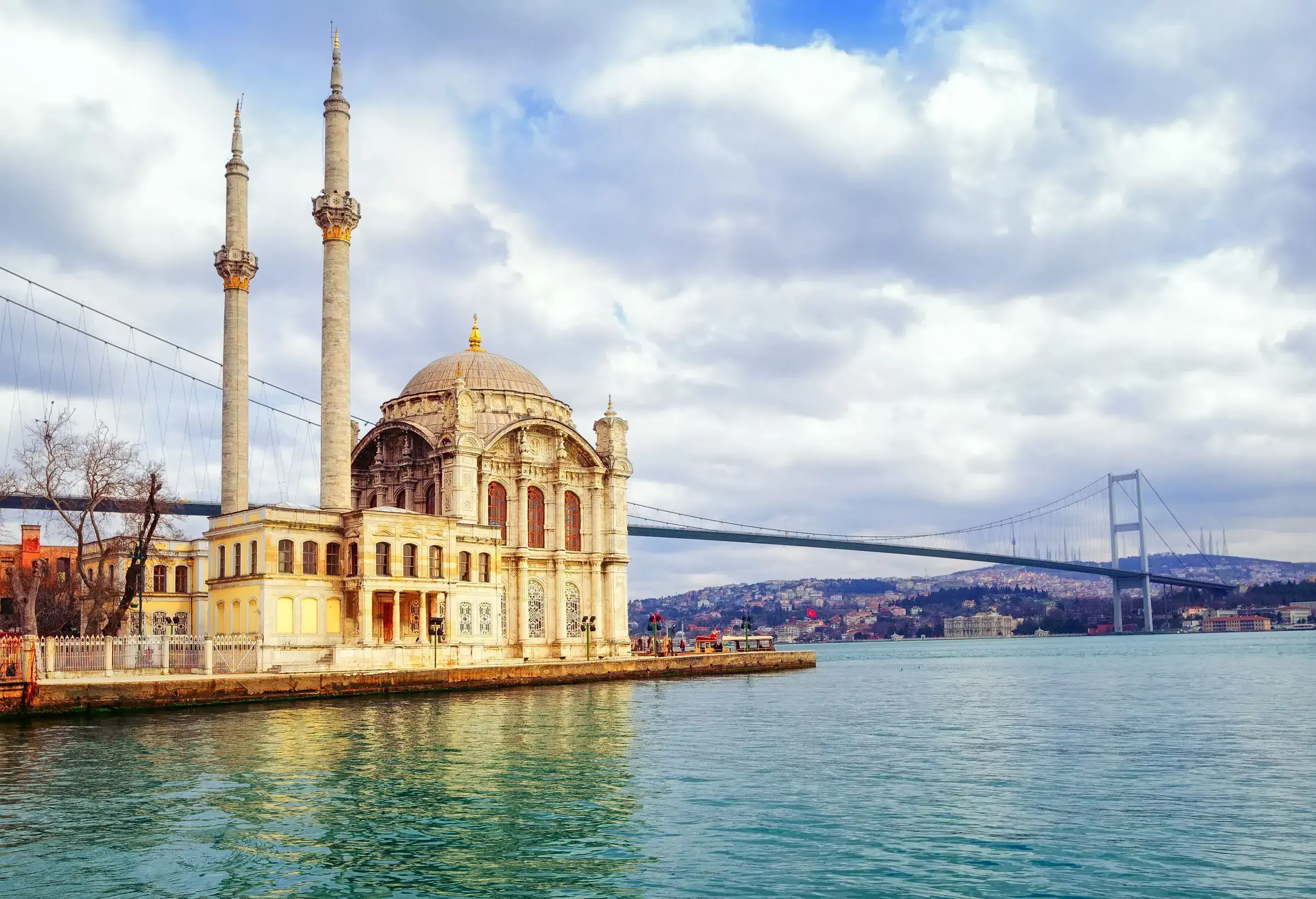 A mosque with slim twin minarets on a pier square beside a suspension bridge.