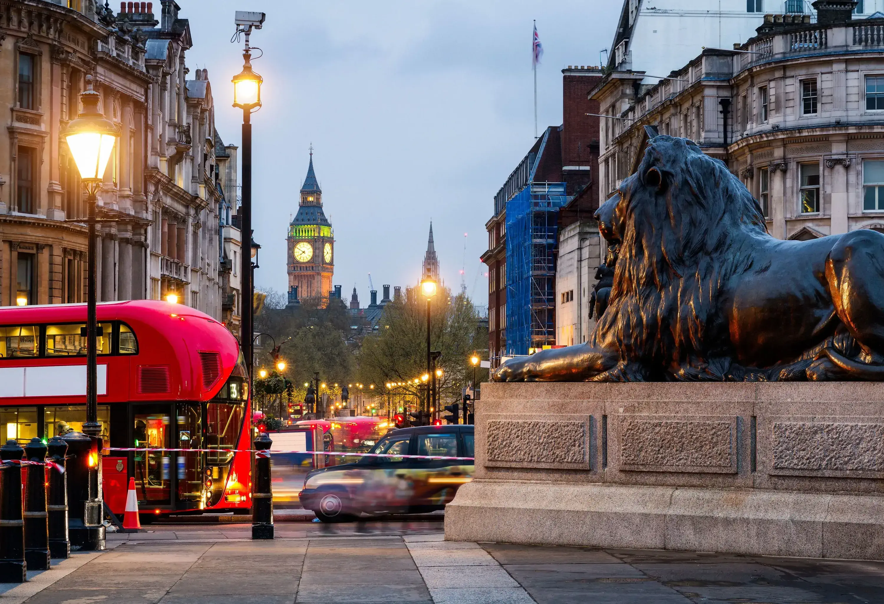 A public square at dusk, showing a prominent lion statue, a red bus and a distant clock tower.