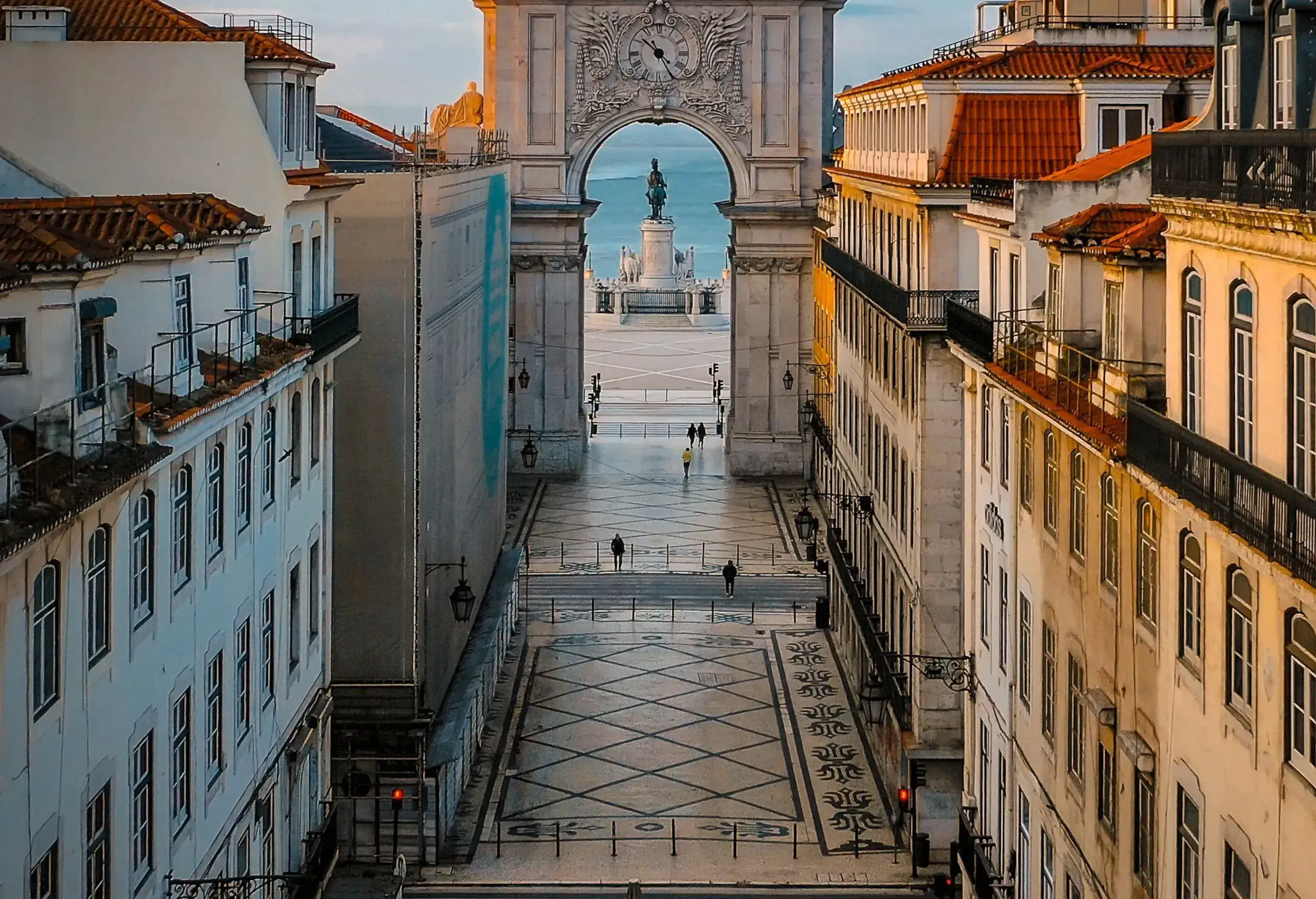 A long street leading to a grand triumphal arch, with classical buildings lining both sides and a large plaza visible beyond the arch.