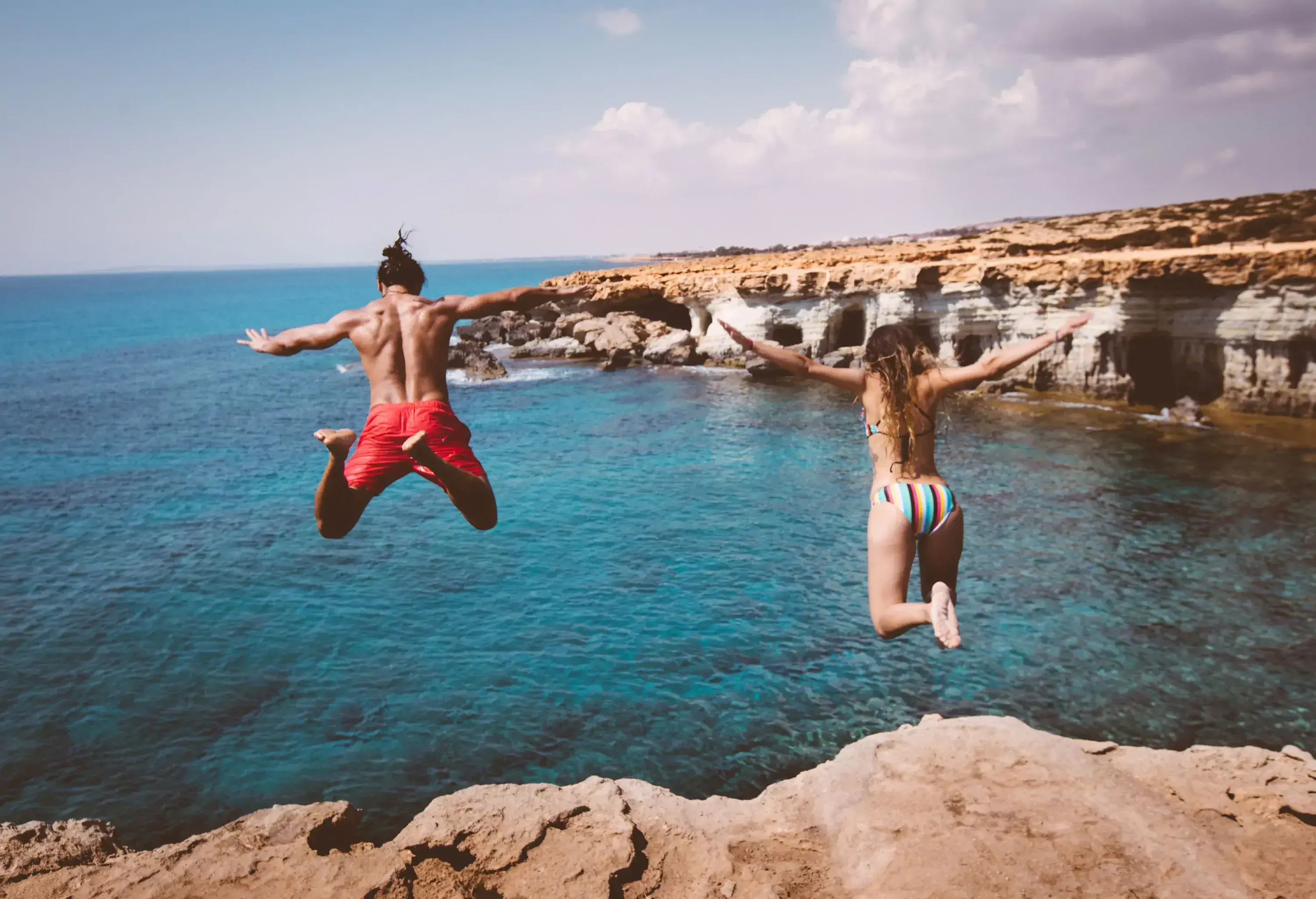 Two people jumping into the azure ocean from a rock ledge.