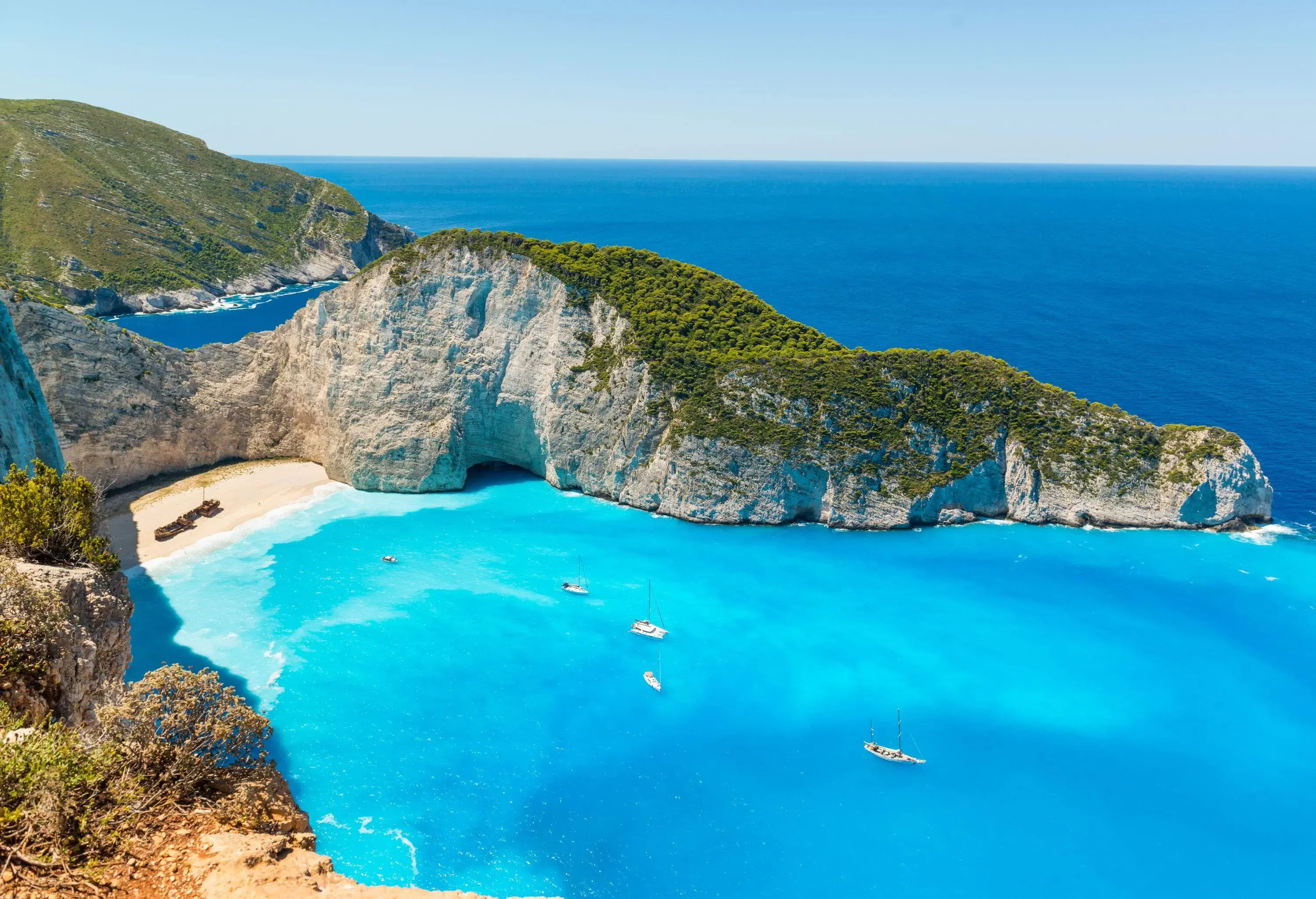 A small cove with azure waters and cruising boats along a white and steep cliff covered in vegetation.