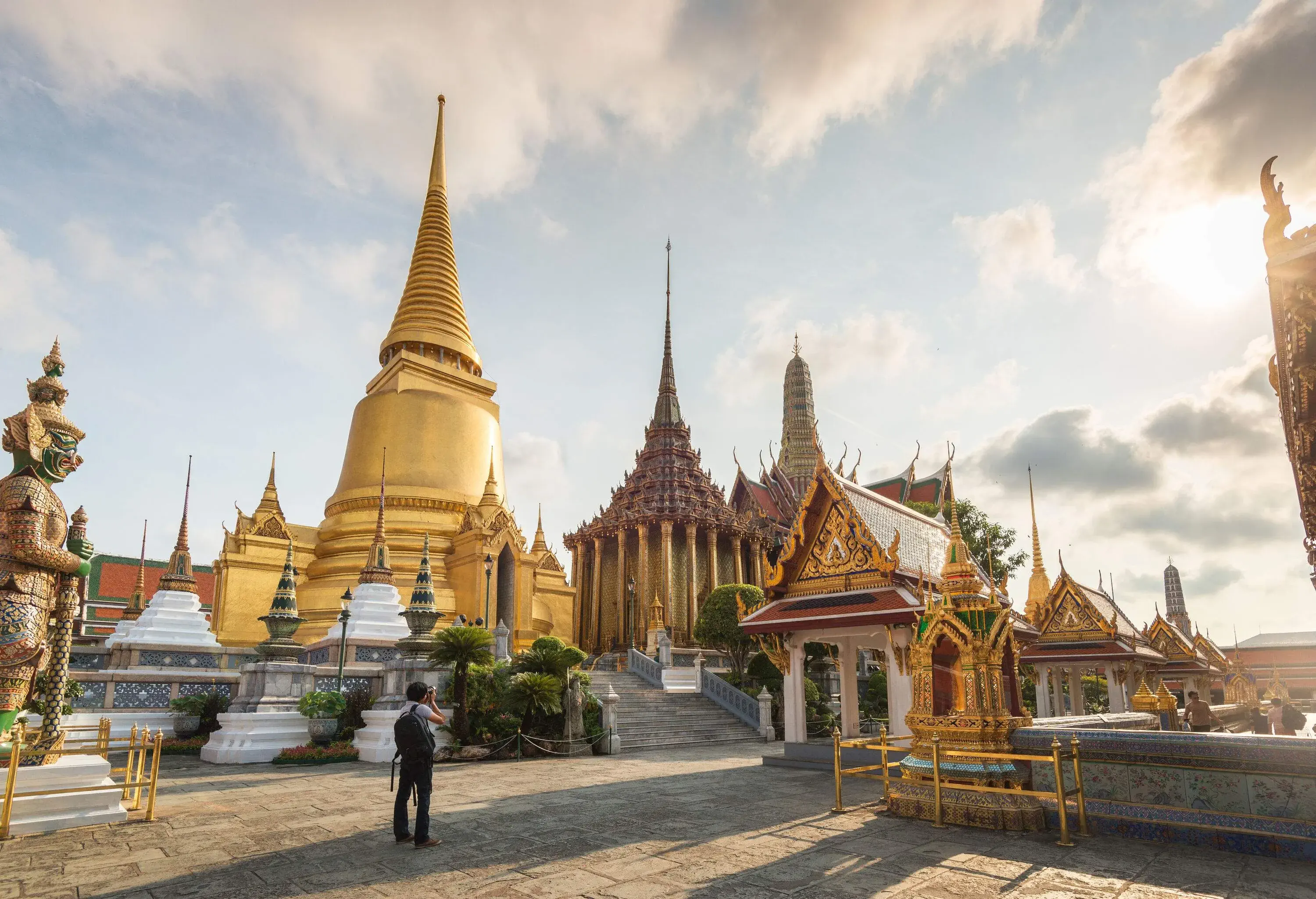A tourist taking photos of the temples inside the Golden Palace with a golden bell-shaped stupa glistening from the sun's rays.