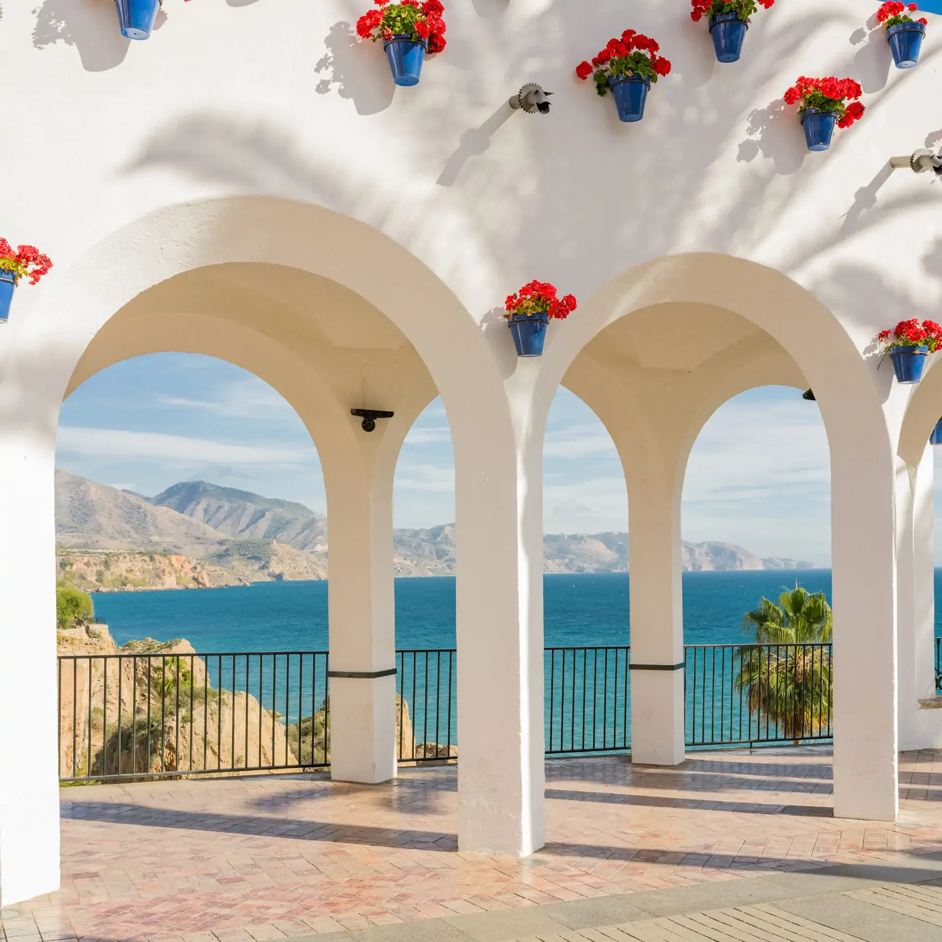 White Mediterranean arches adorned with red flowerpots look out over a calm, blue coastal sea view.