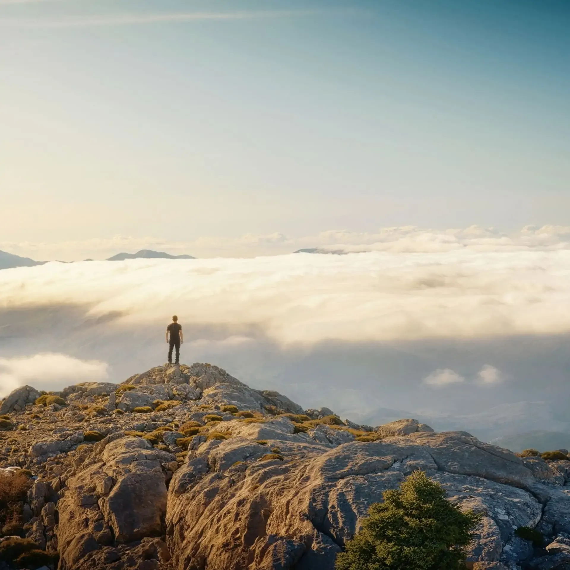 A hiker stands on a rocky mountain peak overlooking a vast sea of clouds under a clear sky.