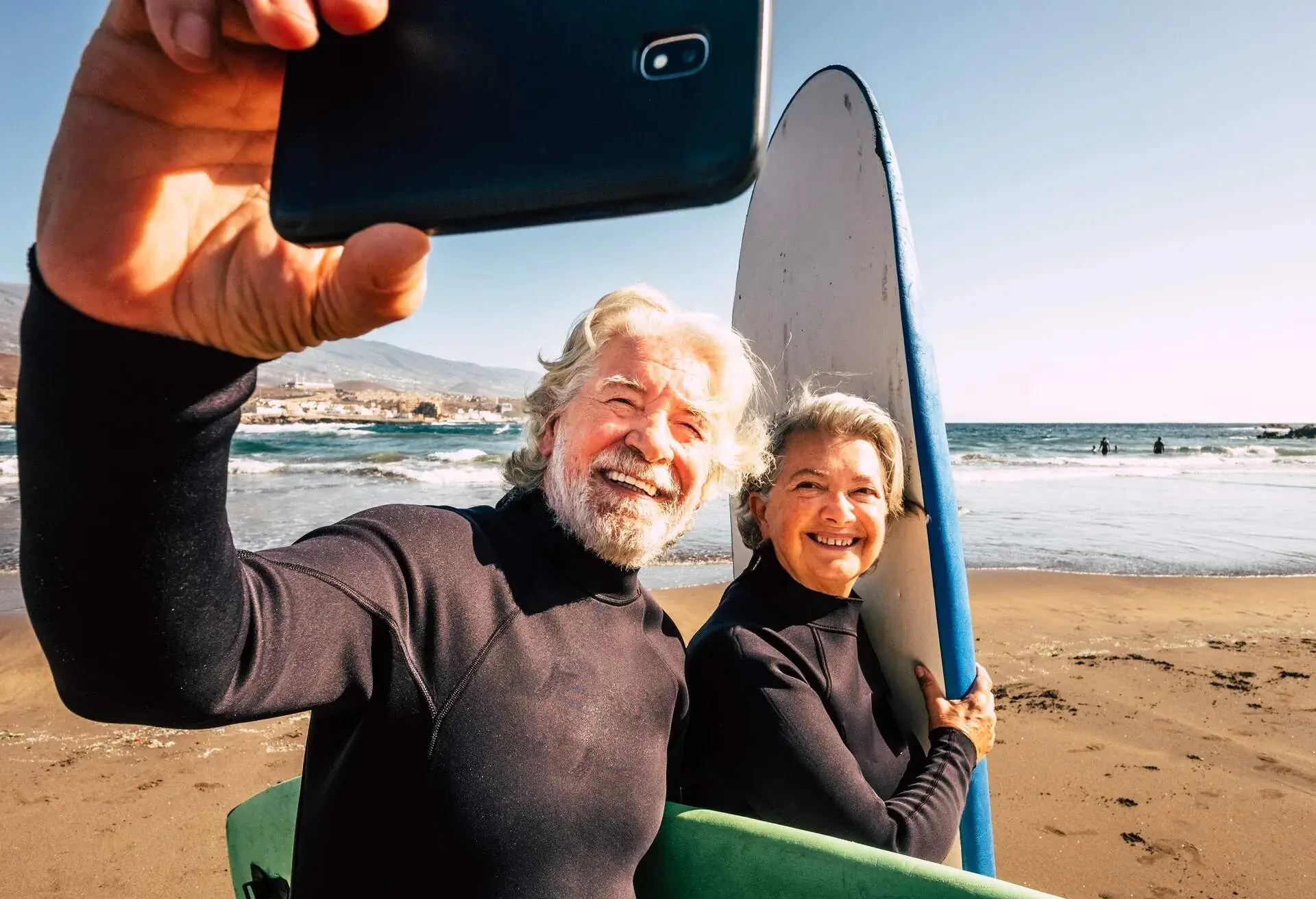 couple of pensioners seniors taking a selfie together at the beach with their wetsuits and surfboards - mature people learning surfing