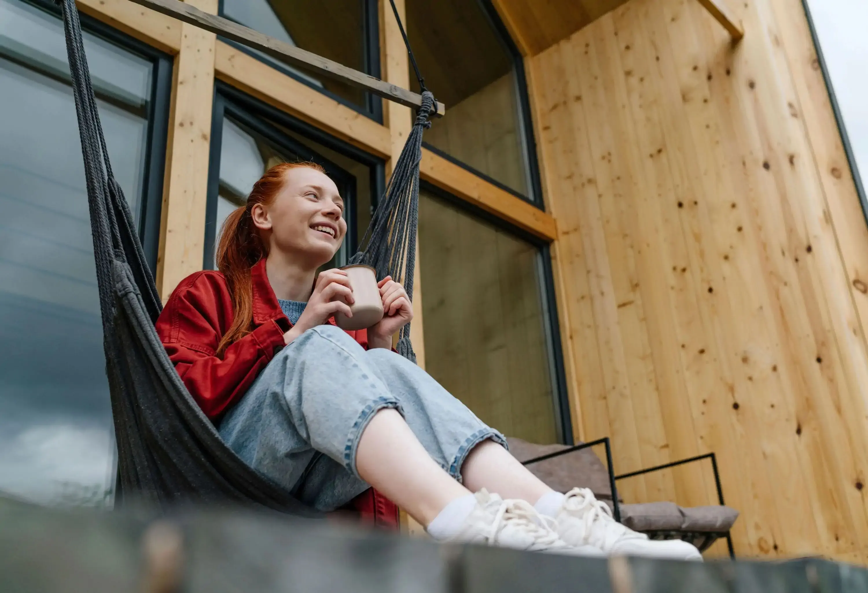 A person sits on a hammock, holding a cup of beverage, smiling as they gaze into the distance, with the rustic charm of a wooden house in the background.