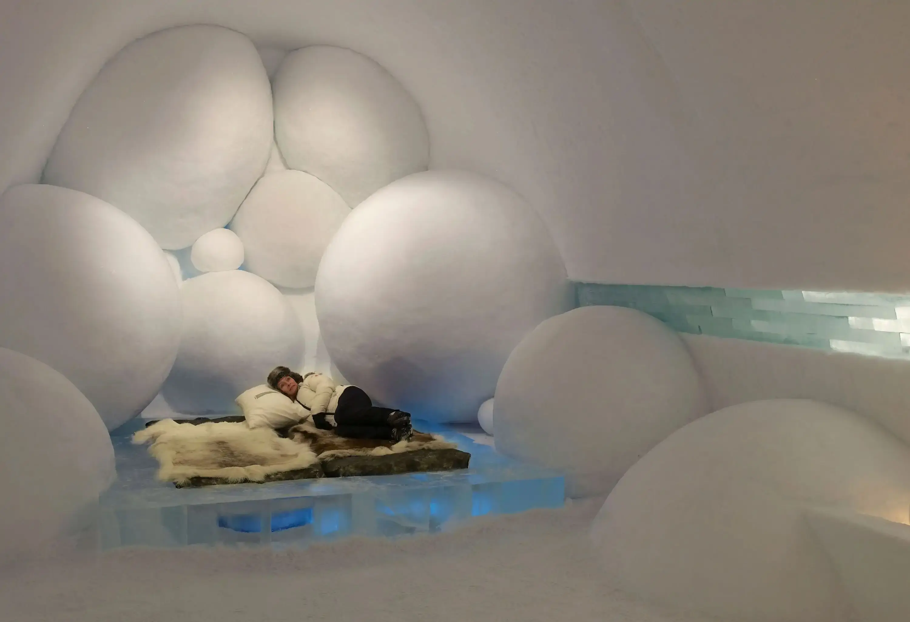 A woman lying down on an animal skin rug in an Icehotel room surrounded by huge snowballs.