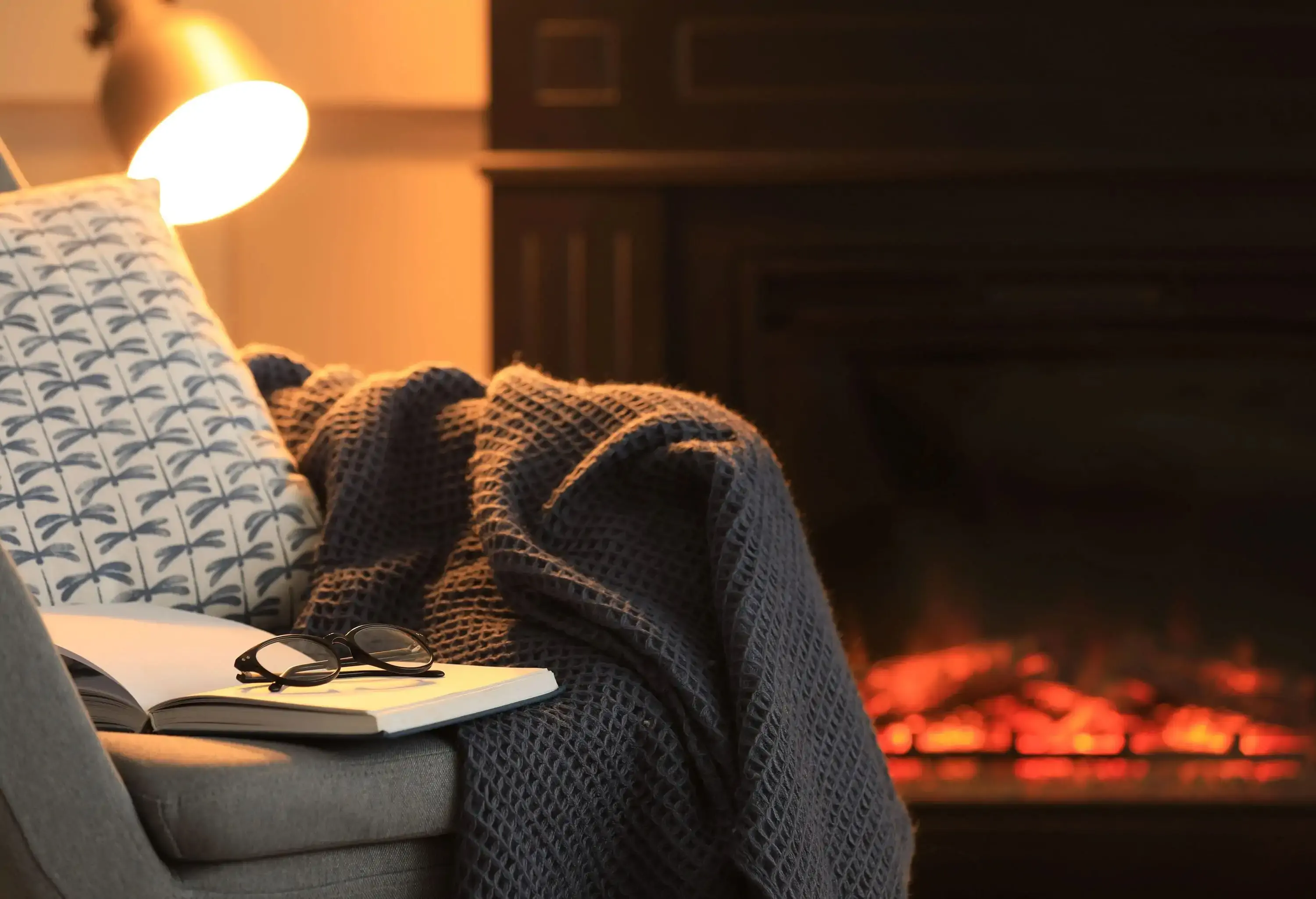 Open book, glasses and plaid on armchair near fireplace