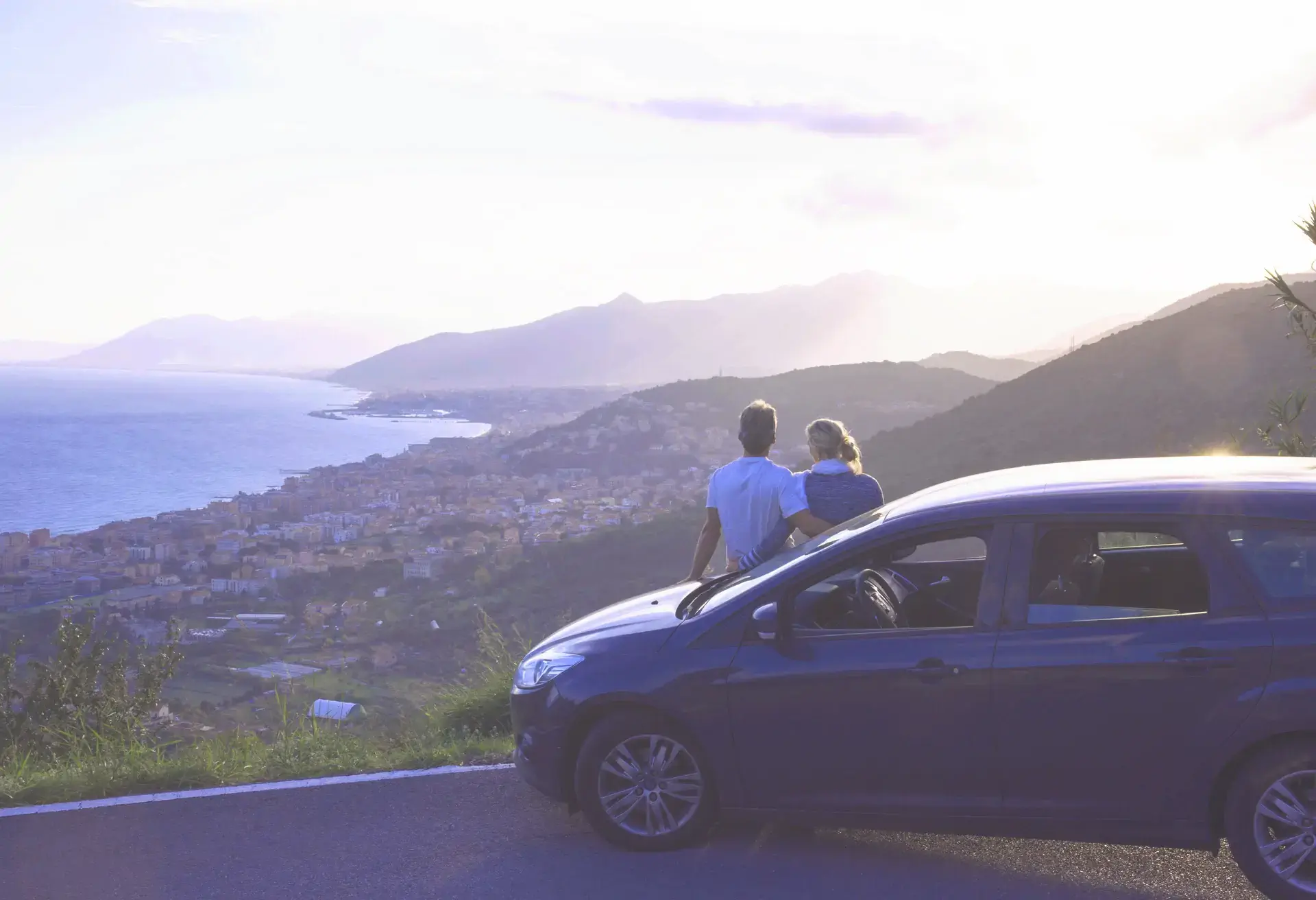 Couple relax against car, look past village to sea