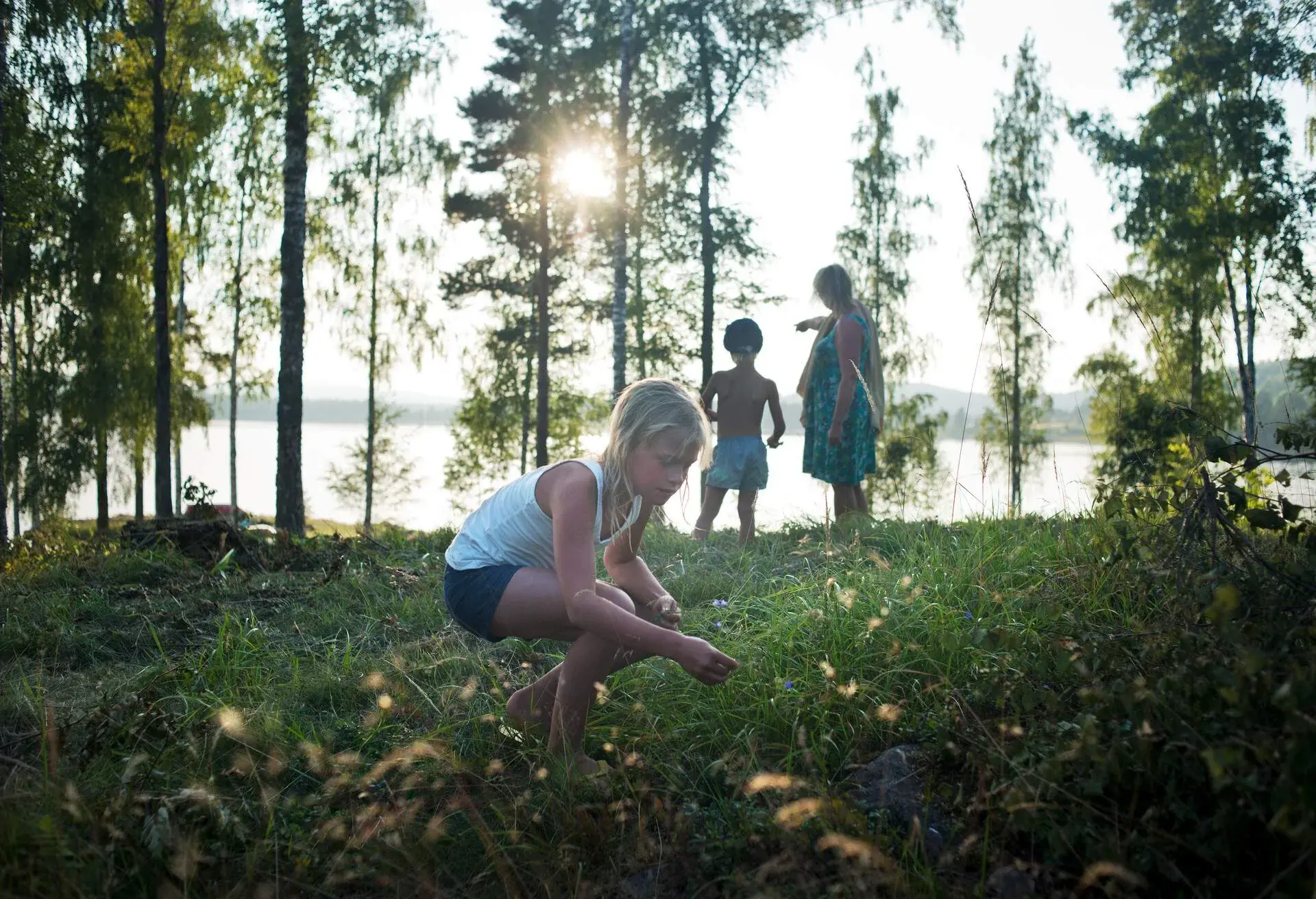 A young girl picking wildflowers in a field with a young boy and a woman standing behind her.
