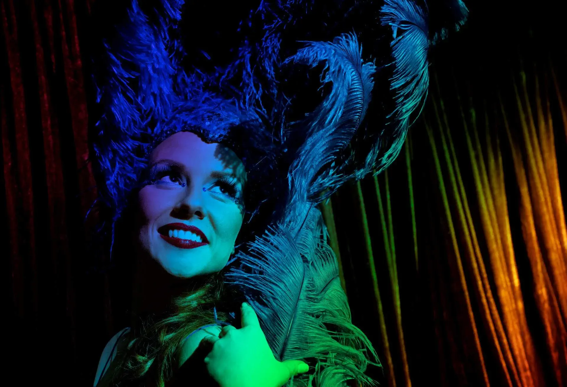 She flashes a seductive grin while standing in front of blue-hued lights and wearing an ostrich feather headpiece.