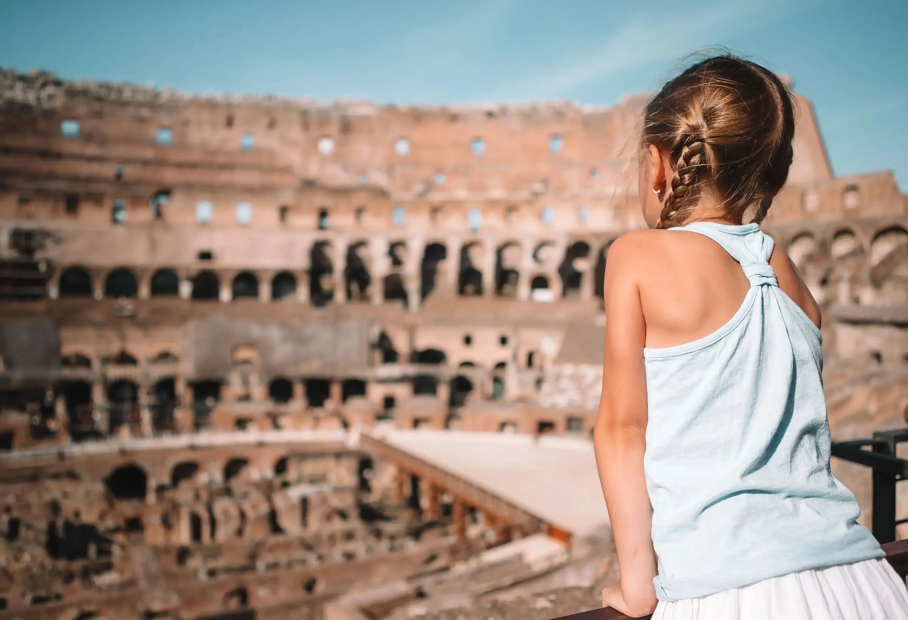 A little blonde girl is standing in an elevated area of the Roman Colosseum, looking at the surrounding.