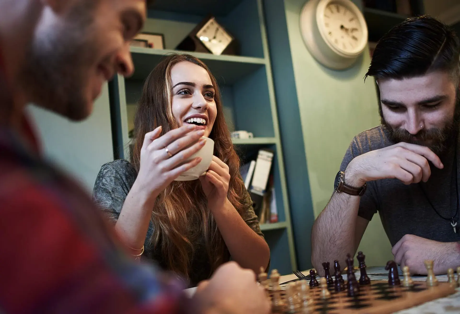 Group of friends playing chess and chatting and laughing in urban cafe. Female holding mug of coffee