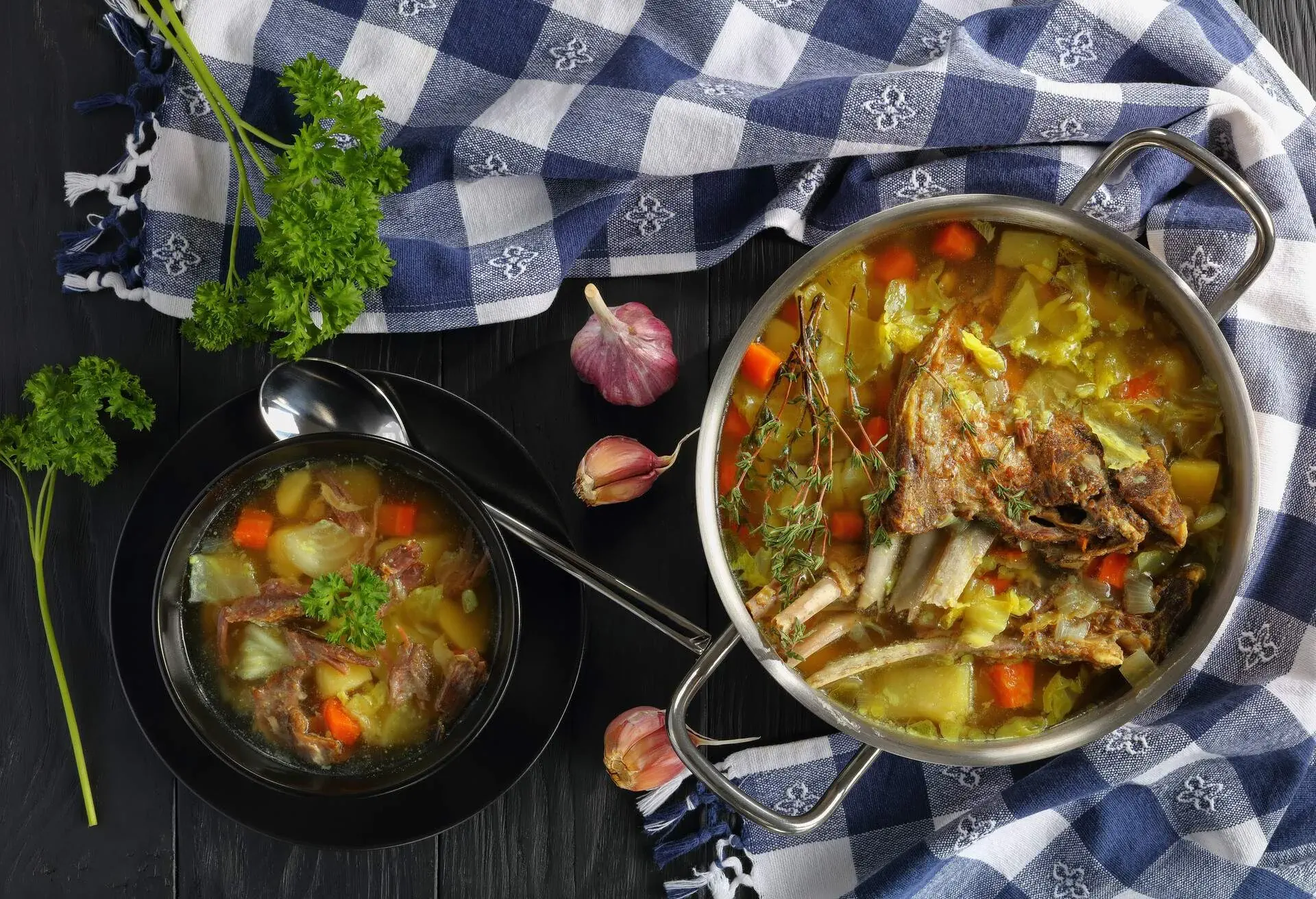 close-up of delicious Icelandic Lamb Soup with vegetables and spices or kjotsupa in a stainless steel casserole pan and in bowl on wooden table with kitchen towel, traditional recipe, view from above