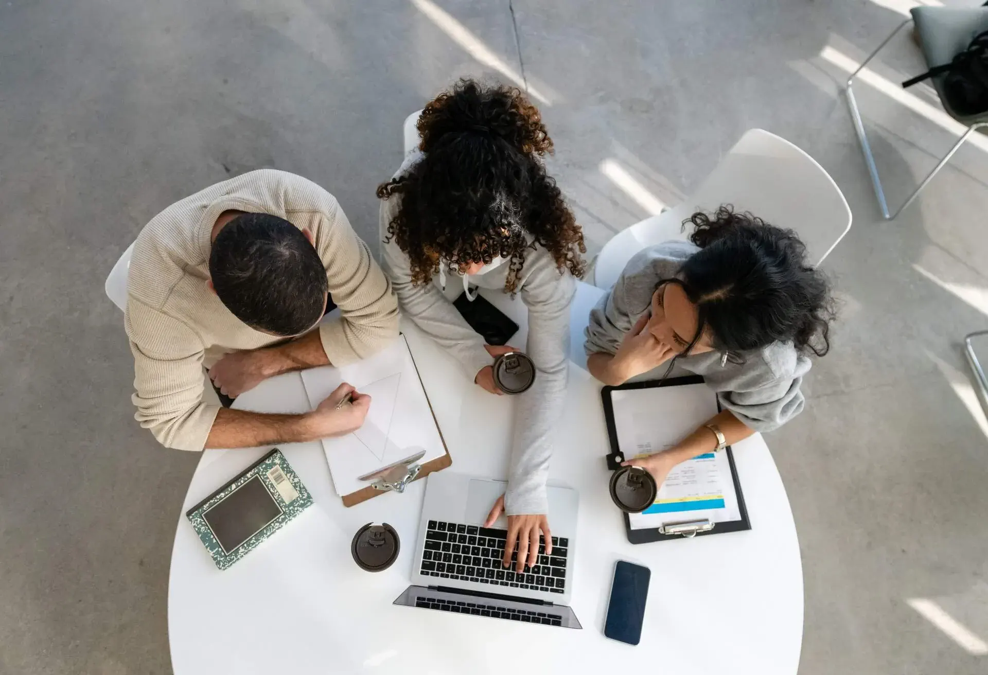 A group of three (3) diverse friends work together on a university assignment one afternoon. The perspective is taken high up and looking down in this overhead shot. They are brainstorming for a project in their business class.