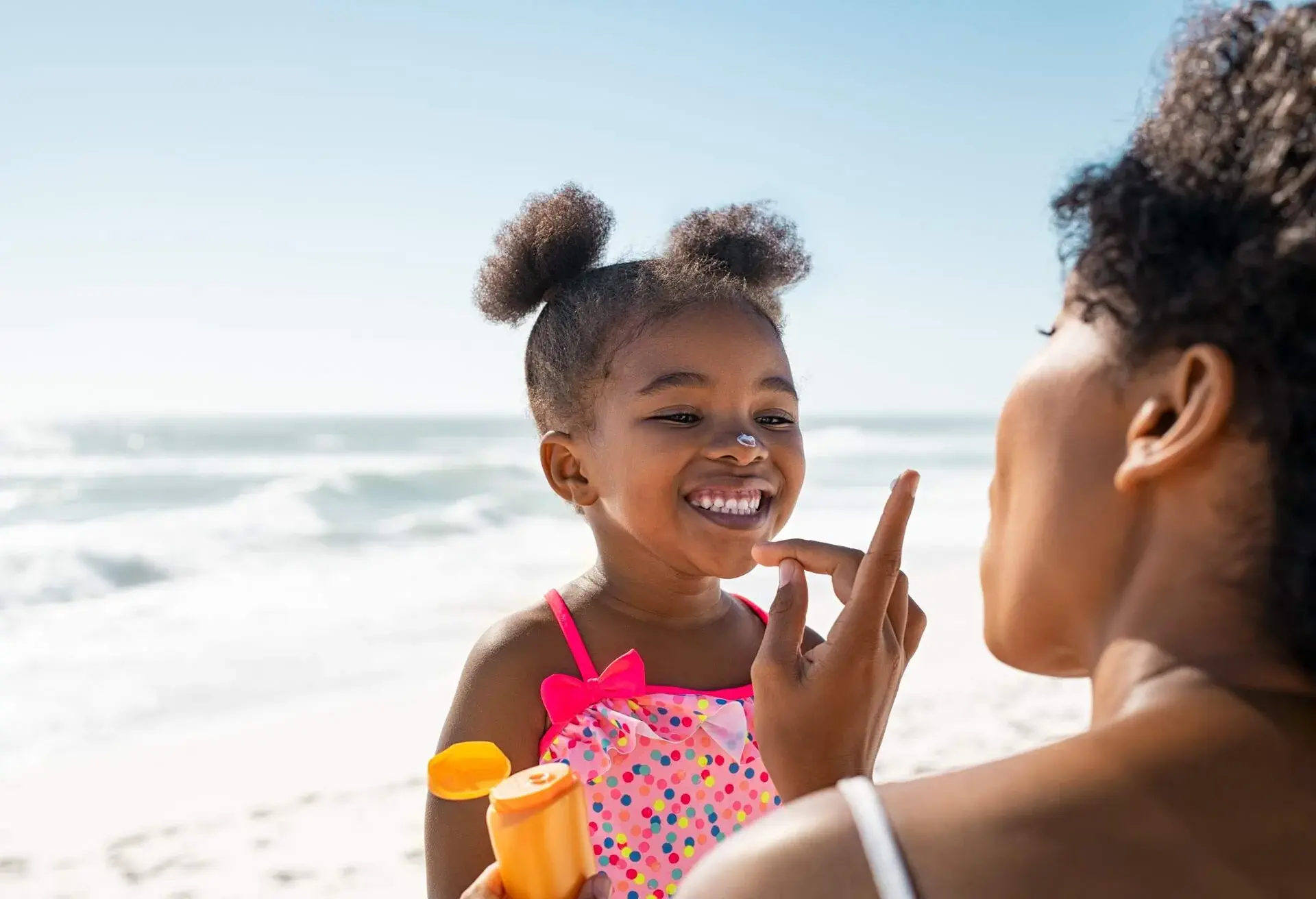 Young mother applying protective sunscreen on daughter nose at beach with copy space. Black woman hand putting sun lotion on female child face. African american cute little girl with sunblock cream at seaside.