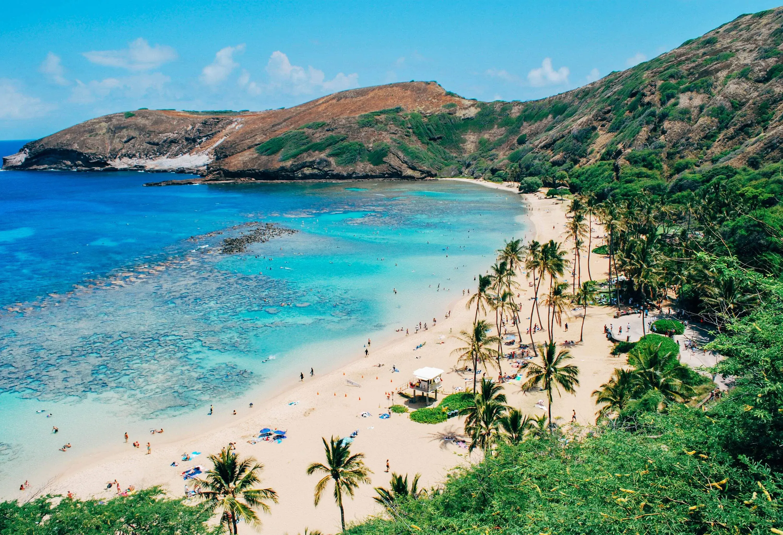 A scenic beach with pristine turquoise water, palm trees, and a lifeguard hut flanked by rugged hills.