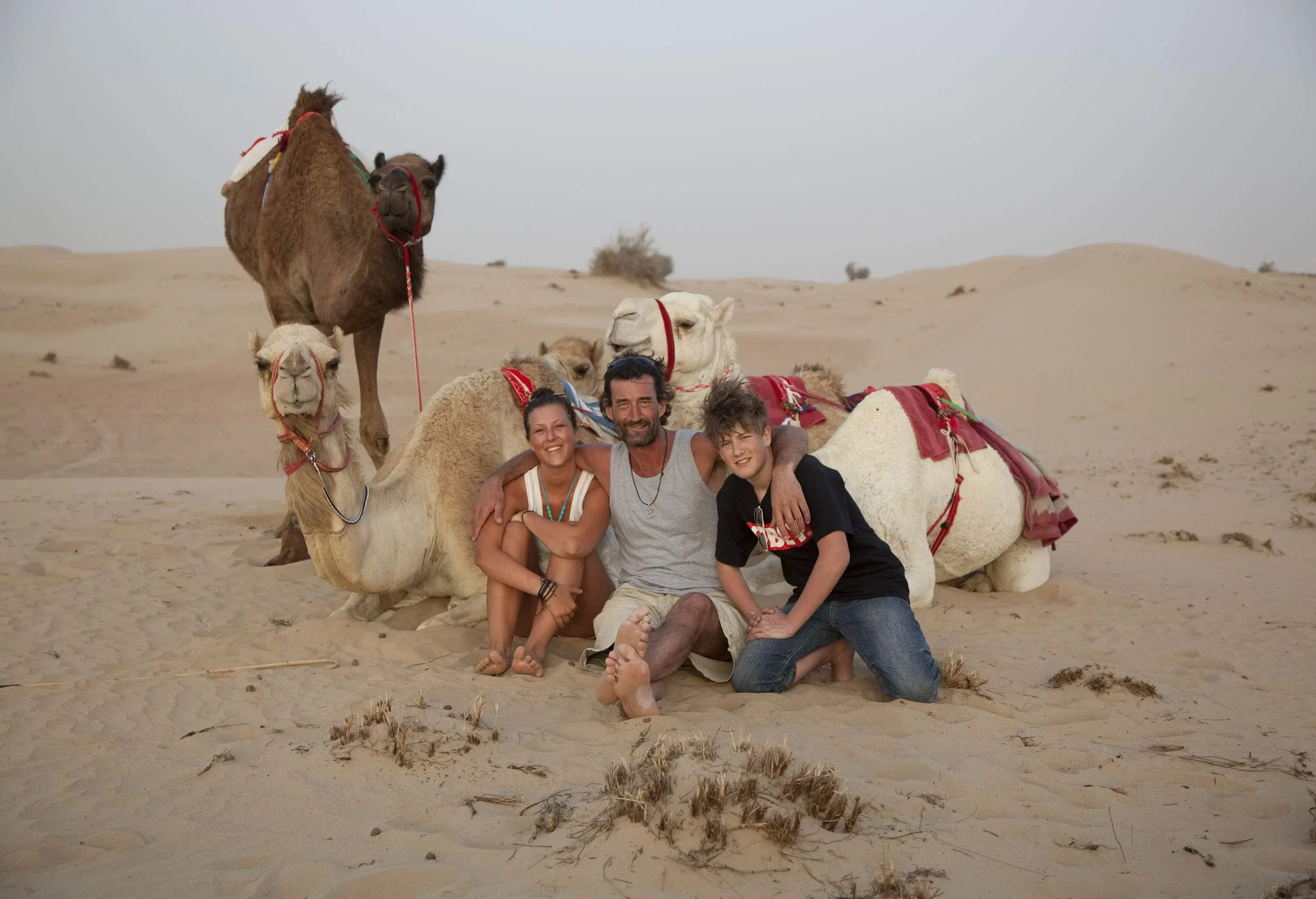 A family of three people posing in front of three camels in the desert.