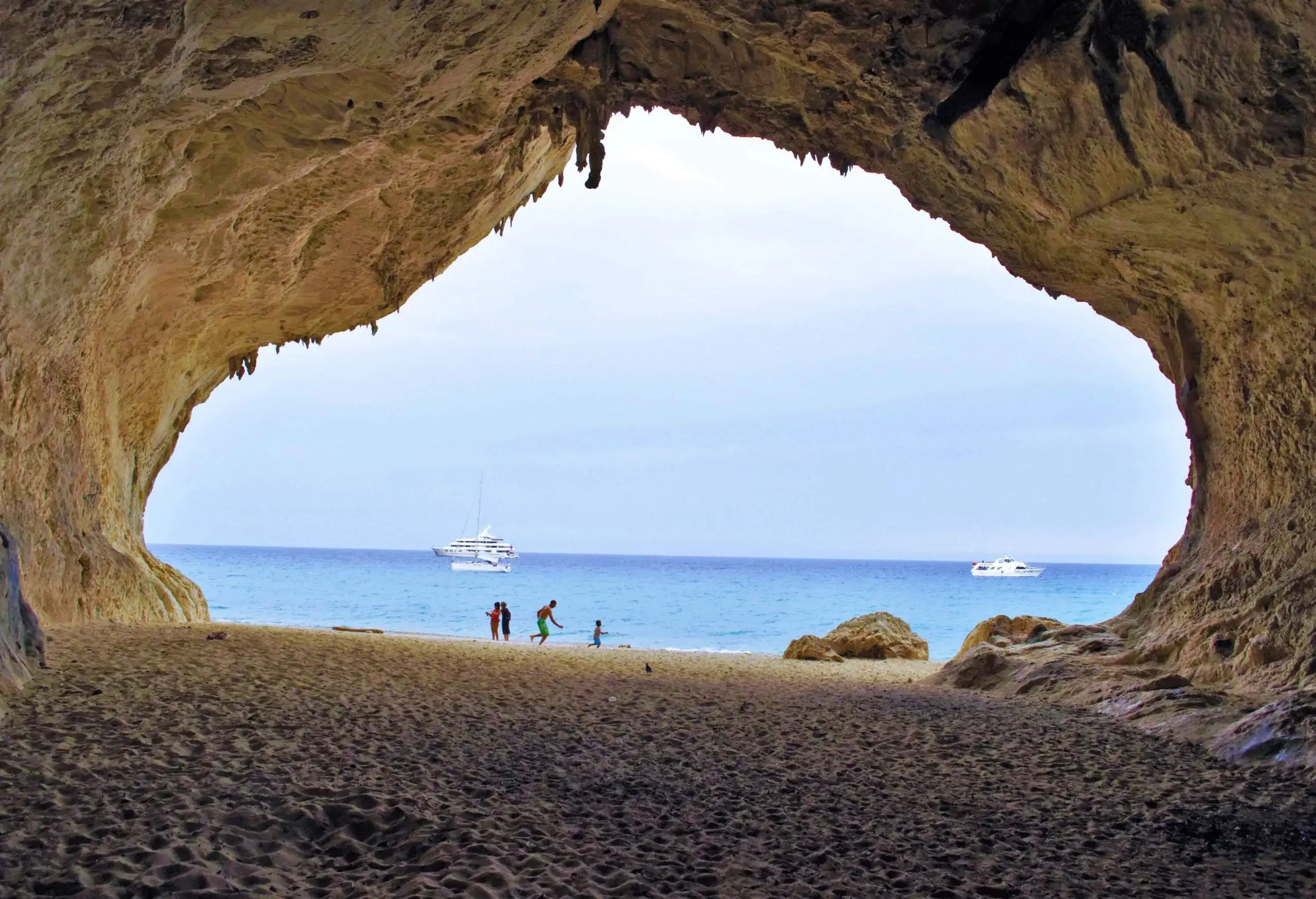 A beach scenery with boats sailing in the distance and people playing on the shore framed by the keyhole of a cave.