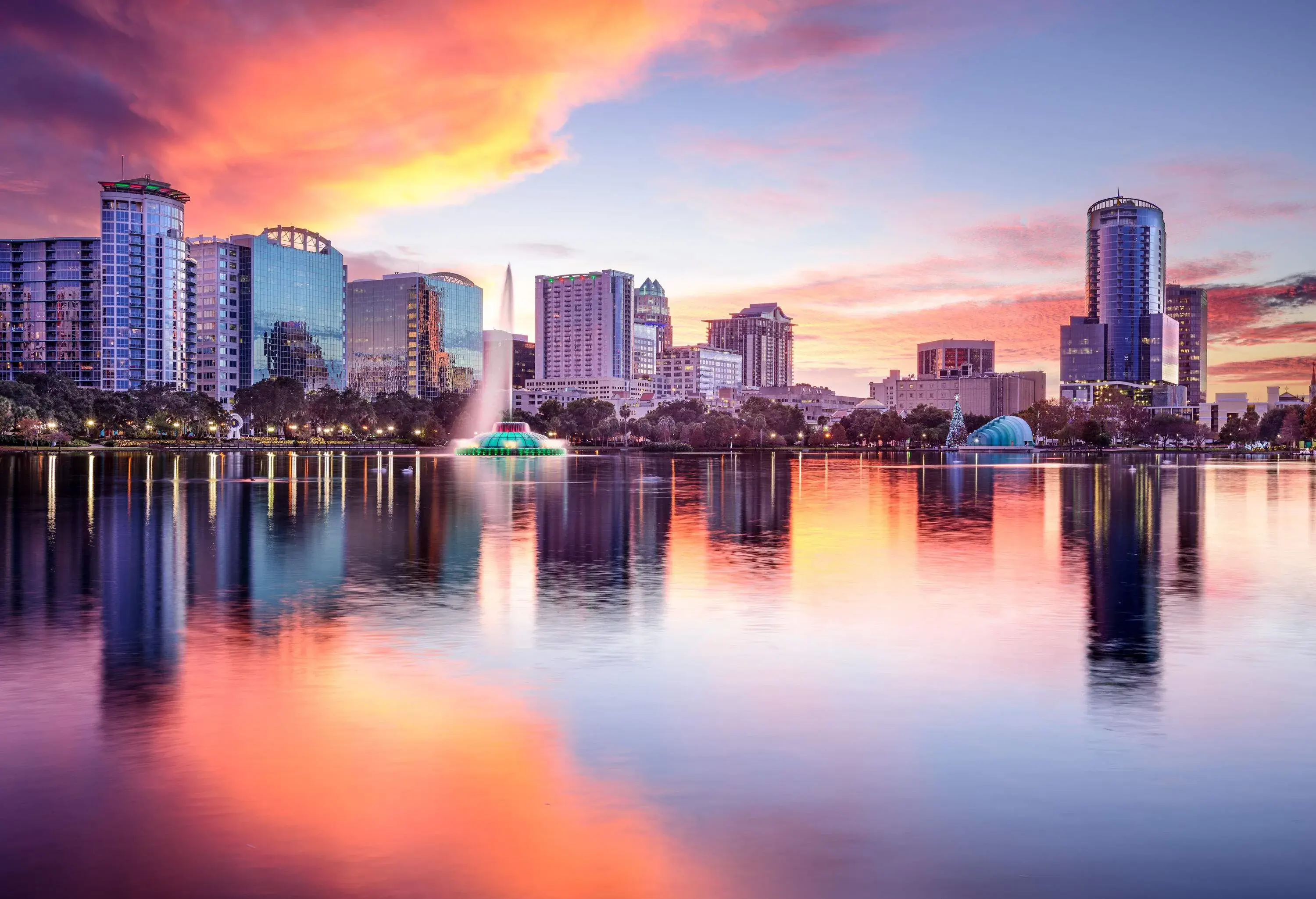 The city skyline is hazily reflected into a peaceful, clear lake that has a fountain close to the shore.