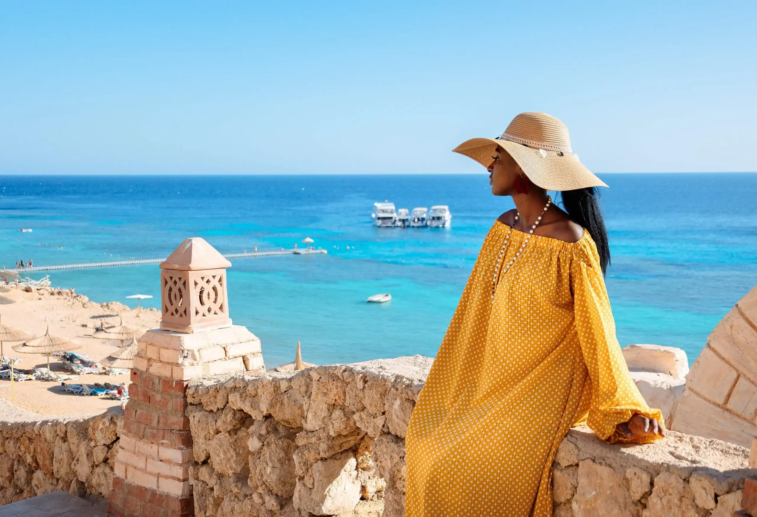 An African lady tourist sits on the stone fence, viewing the protruding dock into the yachts on the turquoise sea.