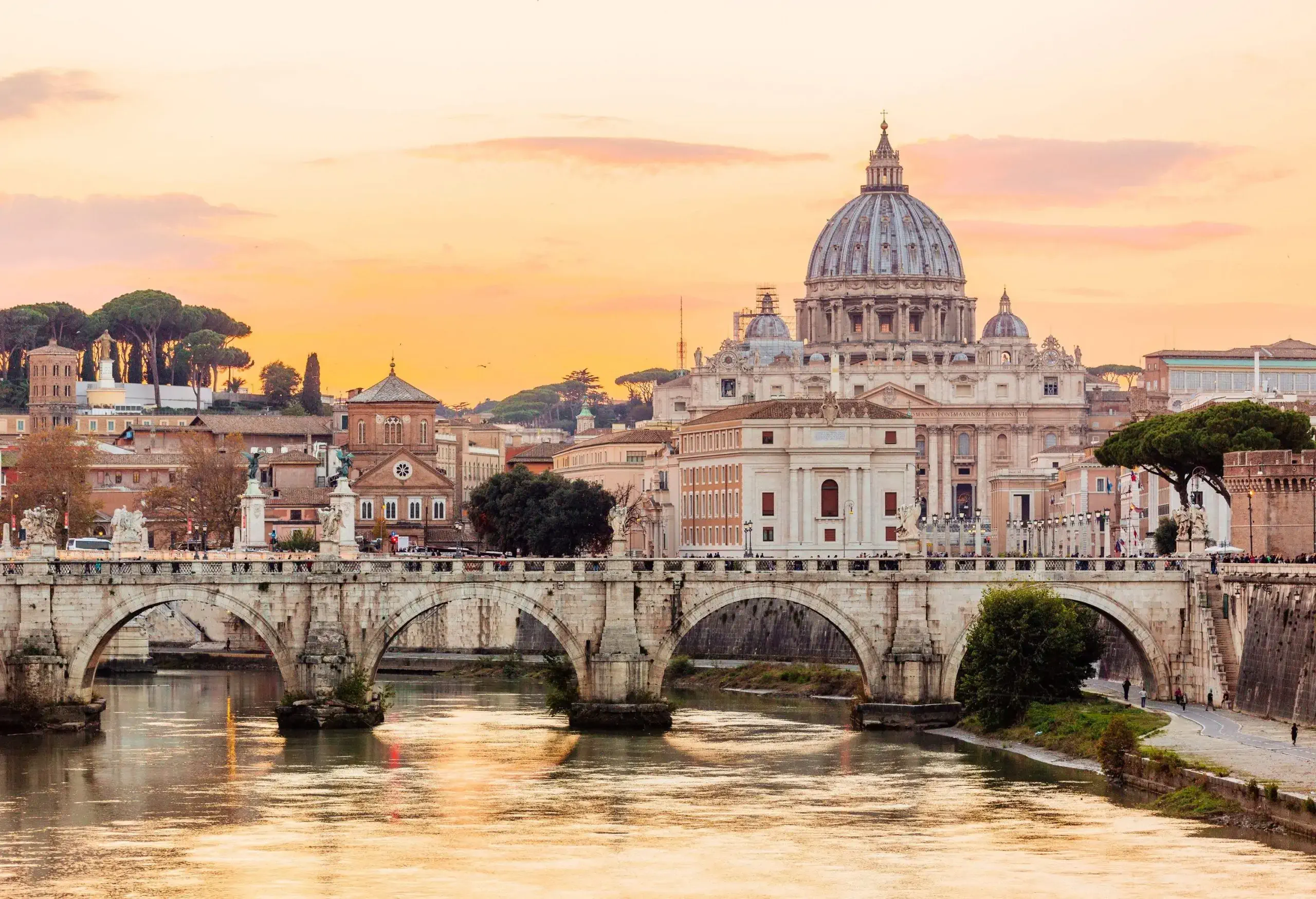 An arch bridge across a calm river with distant views of a domed church rising above a city under an orange sky.