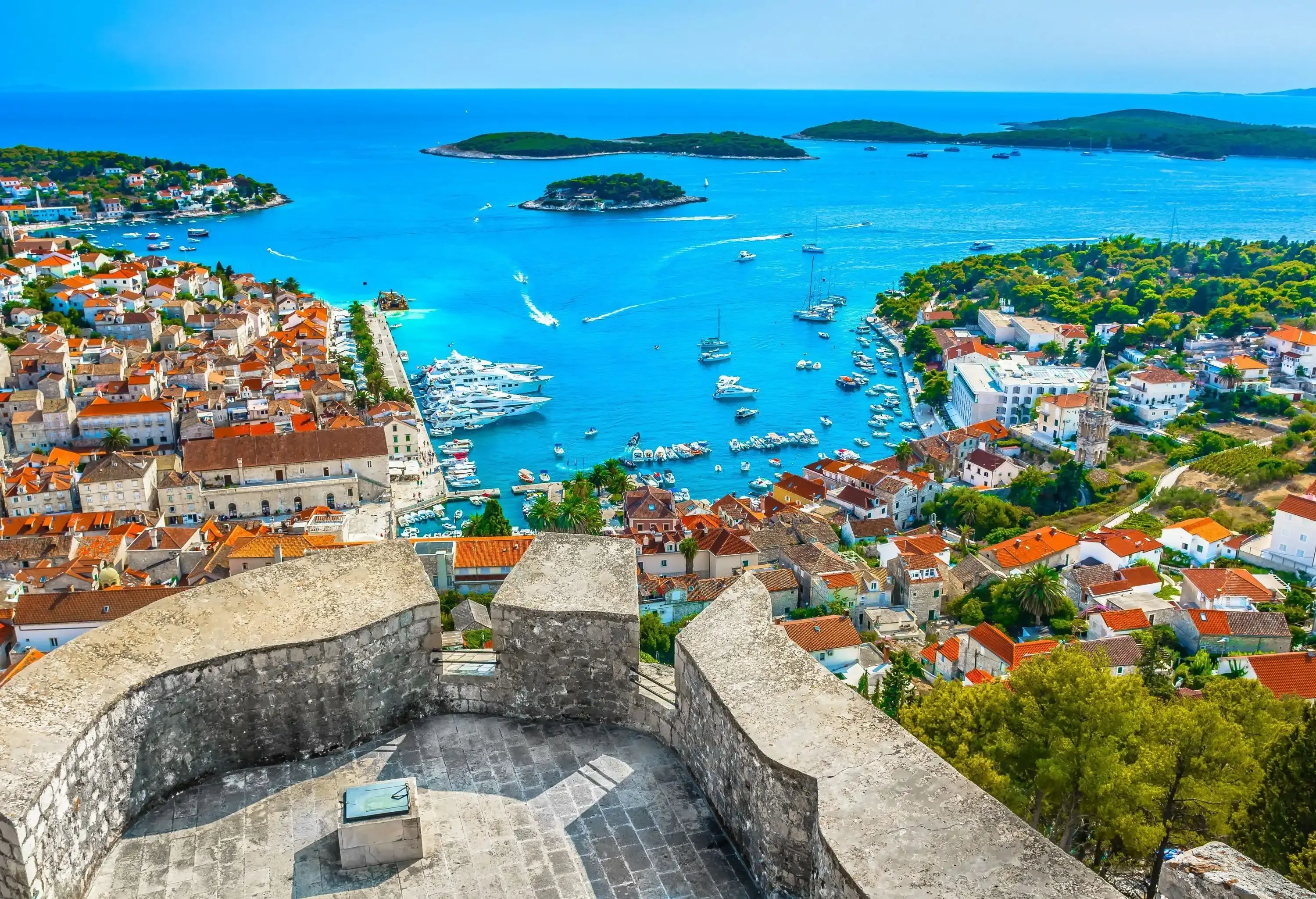 An elevated fortress overlooks the calm turquoise sea, surrounded by clusters of houses and dotted with small forested islands.