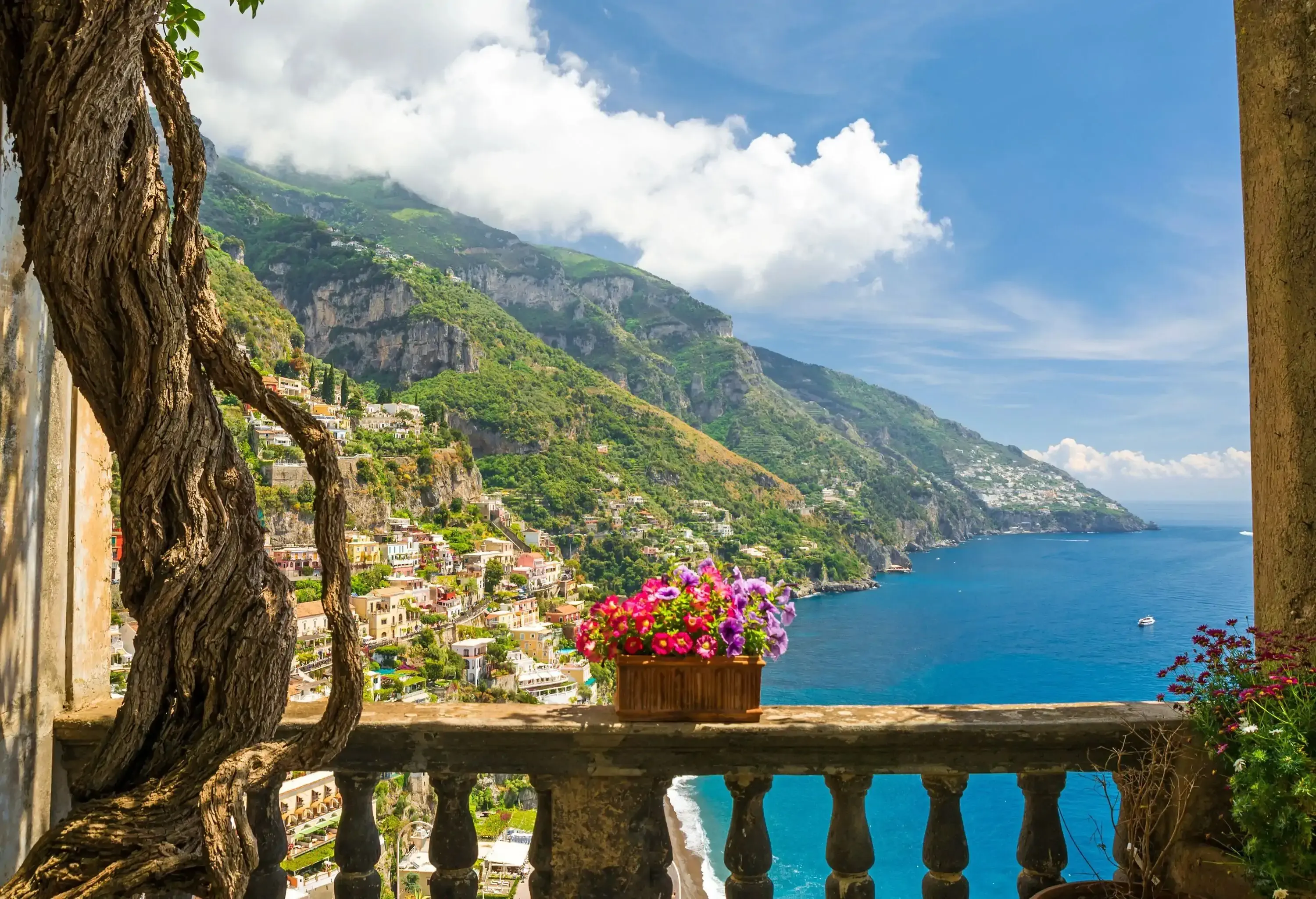 A balcony with flowers with a view of houses on the steep mountainside overlooking the blue sea.