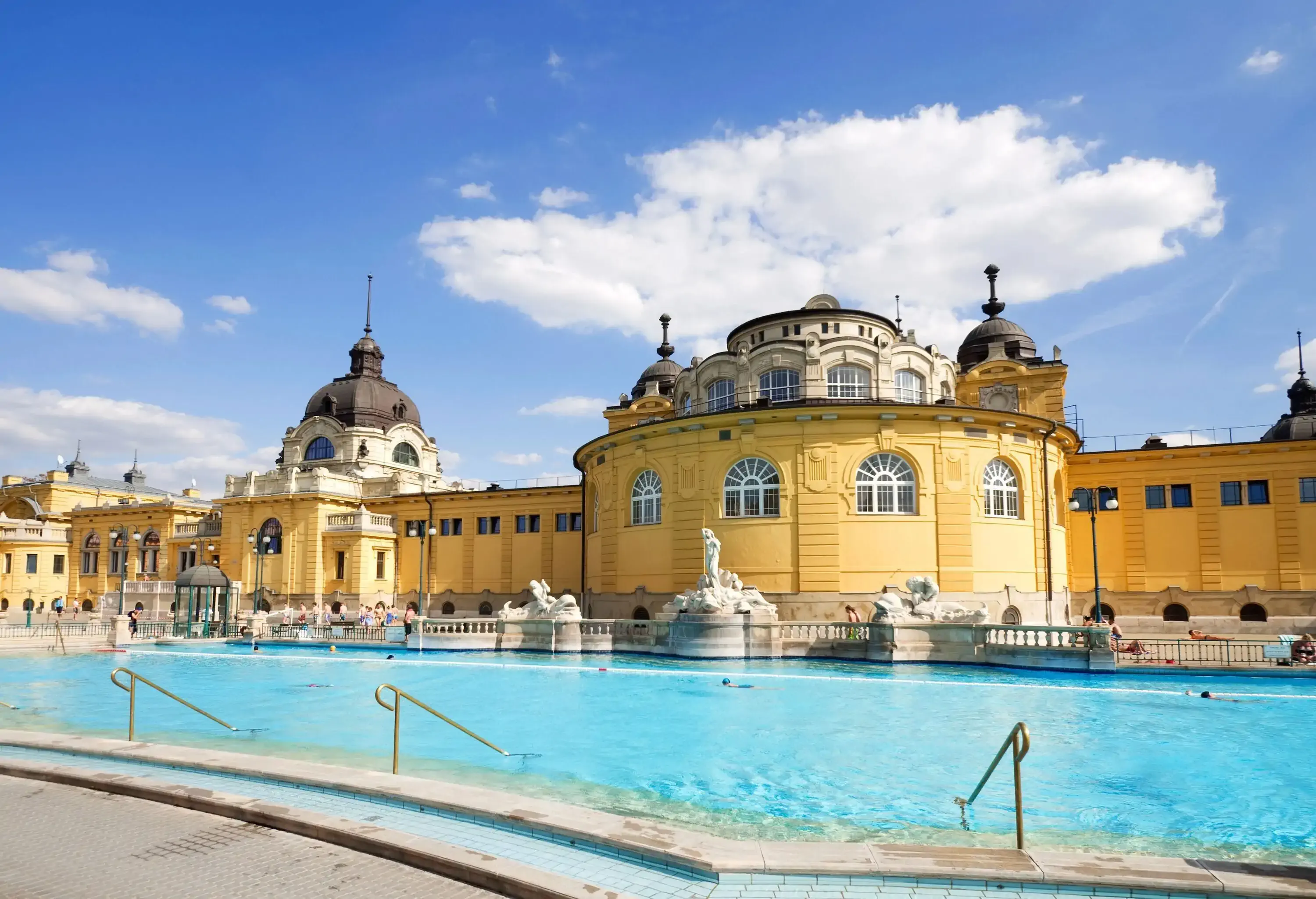A pool of clear water in a neo-baroque thermal complex with cupolas, yellow facades, arched doors and windows.