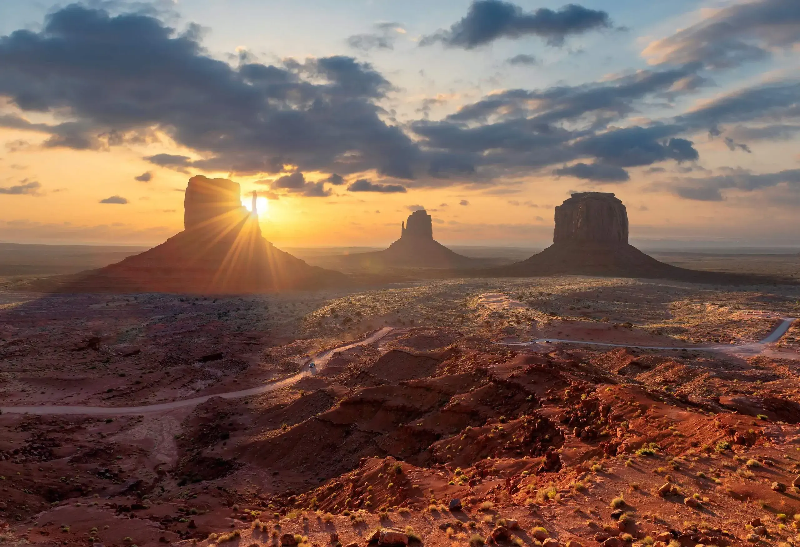 The sun rises in a dramatic sky above three iconic rock formations emerging in the rugged valley.