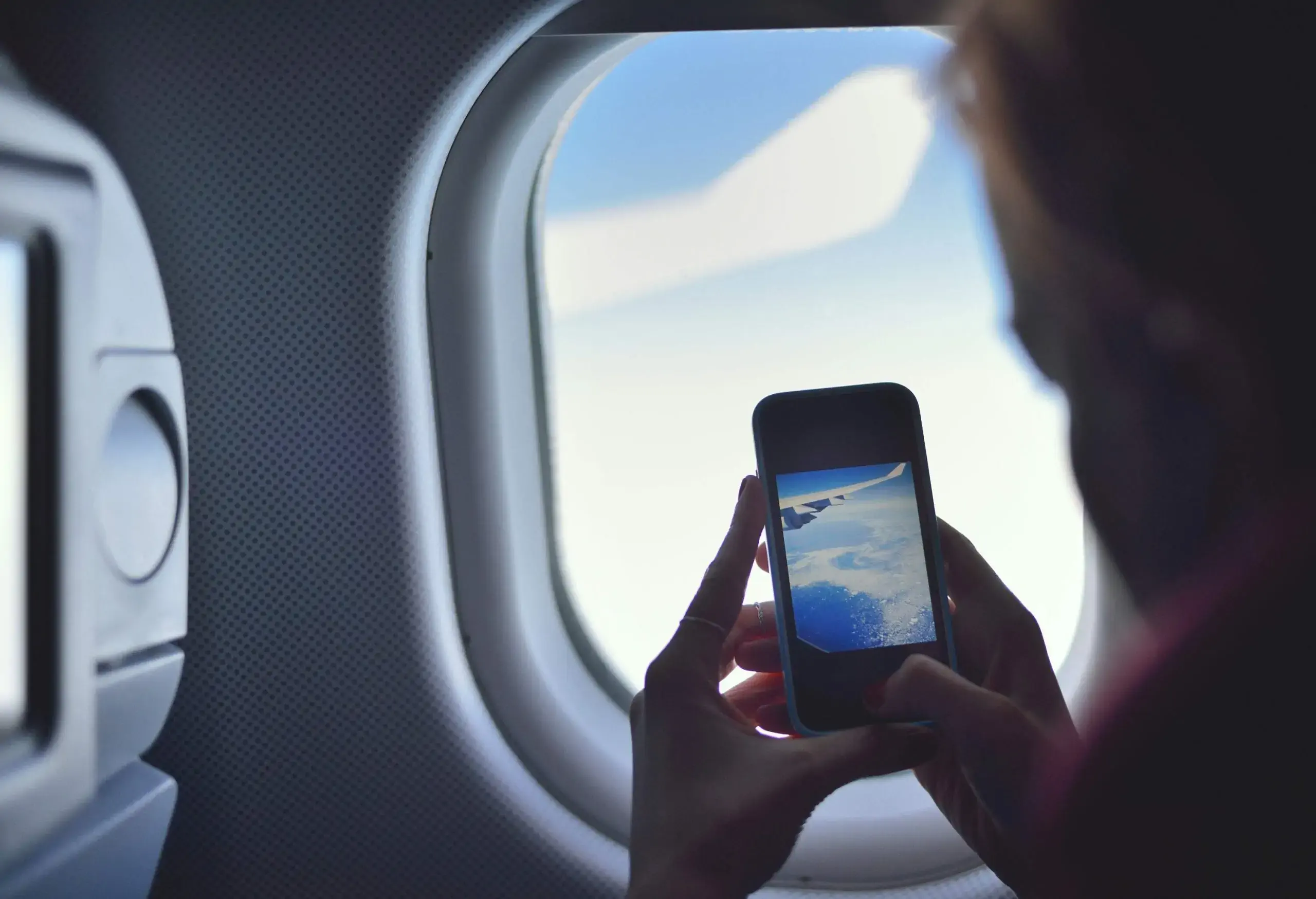 A passenger seated inside an aircraft by the window captures a photo of the white clouds using her smartphone.