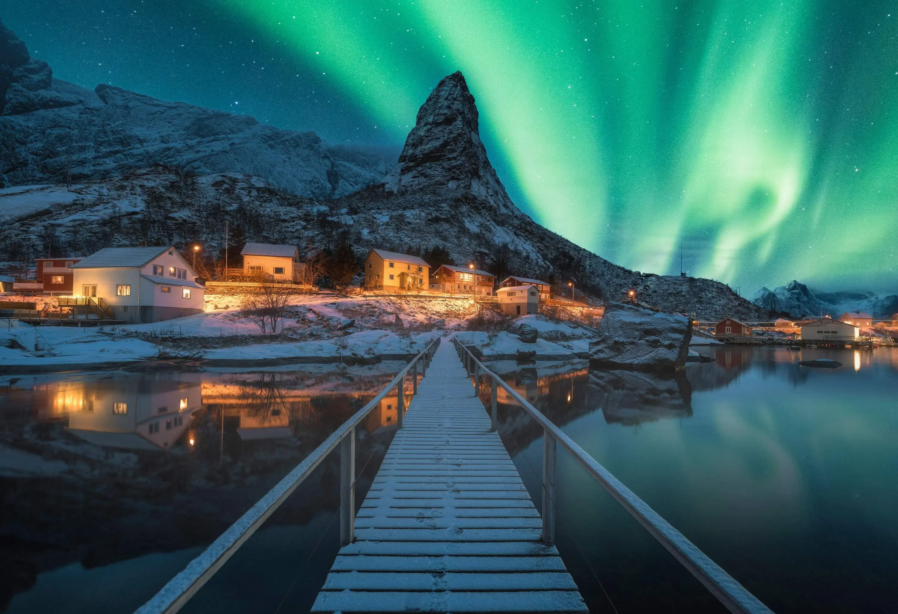 A boardwalk across the sea to a small fishing village lit up by the northern lights blazing across the sky.