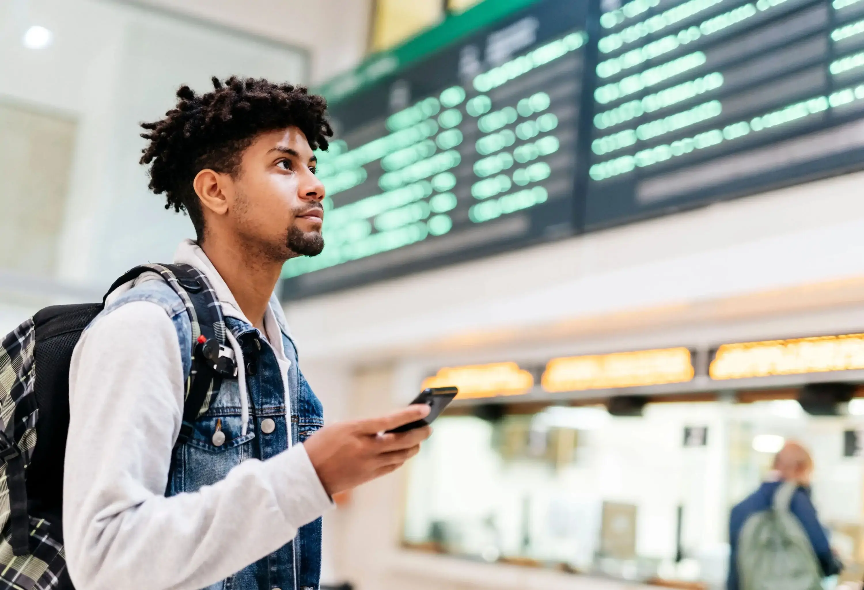 young afro man holding a mobile phone looking at the departures of planes in an airport
