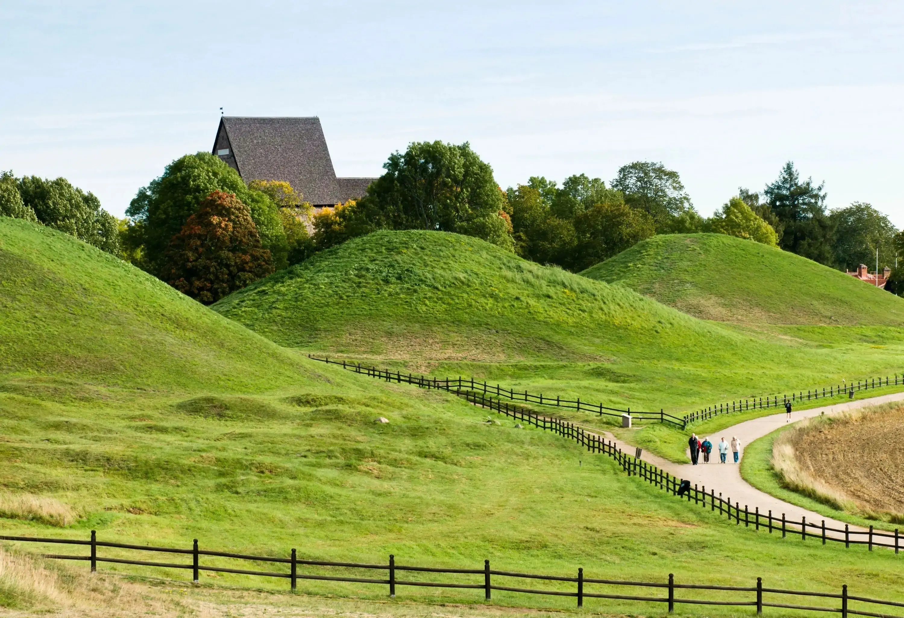Several people walk on a wooden fenced pathway alongside lush green burial mounds surrounded by trees.