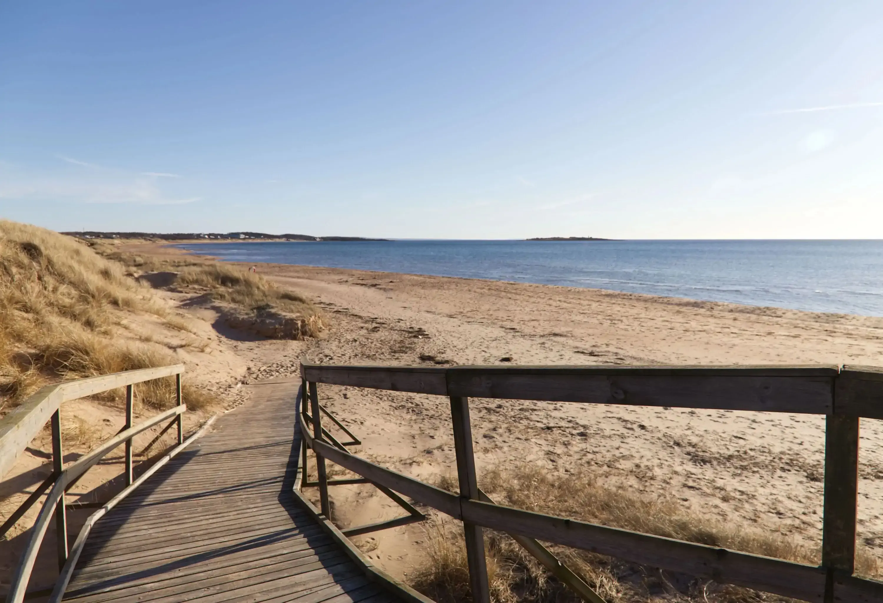 A wooden boardwalk heading down a brown sand beach with calm waters under a clear blue sky.