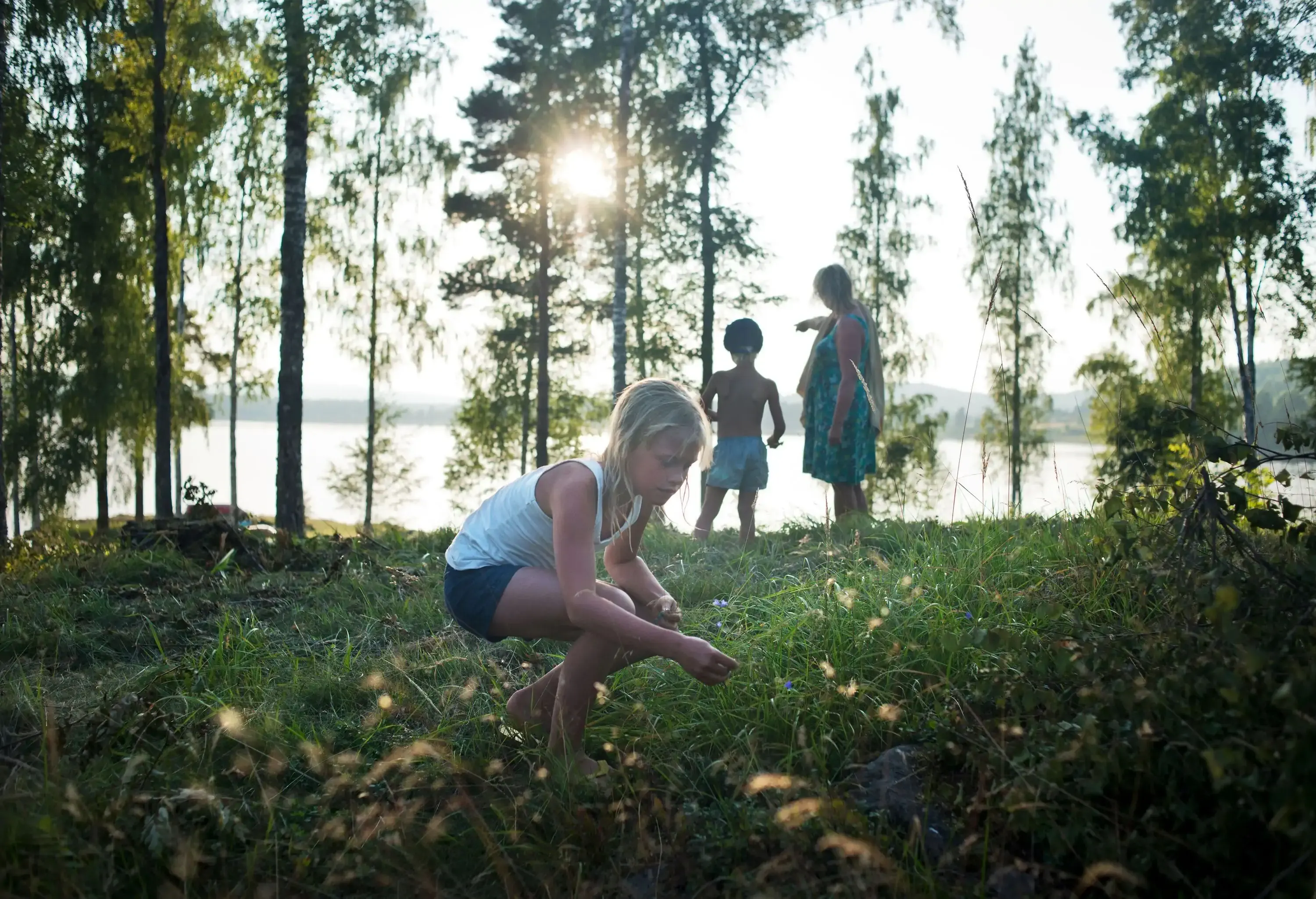 A young girl picking wildflowers in a field with a young boy and a woman standing behind her.