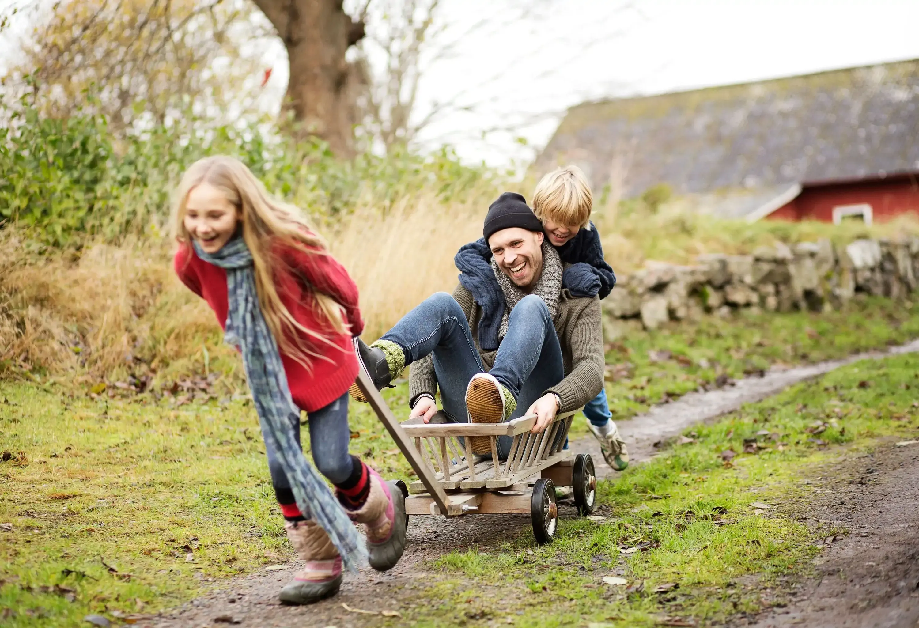 A young girl pulling her father and brother in a wagon on a dirt path.