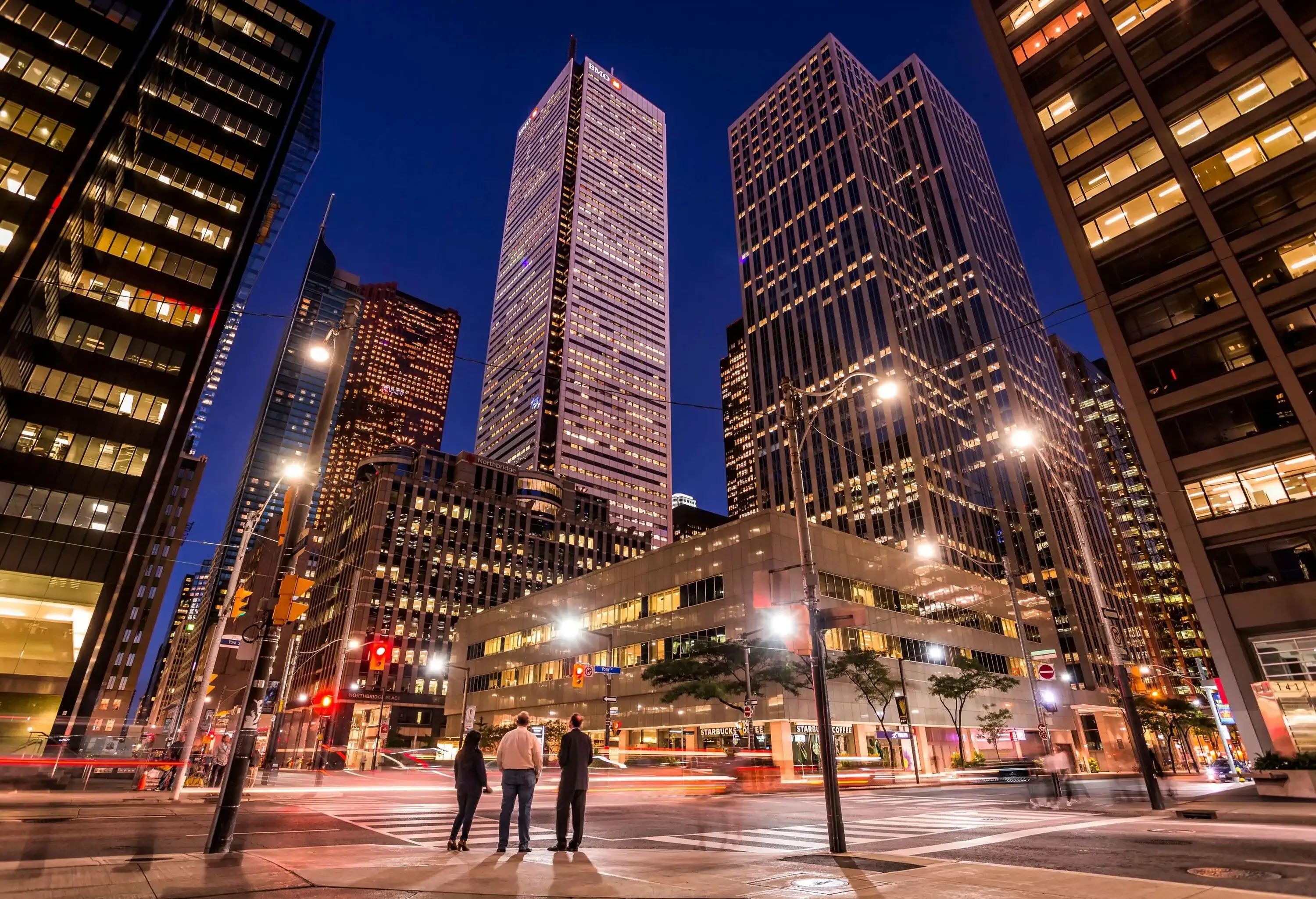 Three people standing along a pedestrian lane in the city centre lit up by the luminous buildings surrounding it.