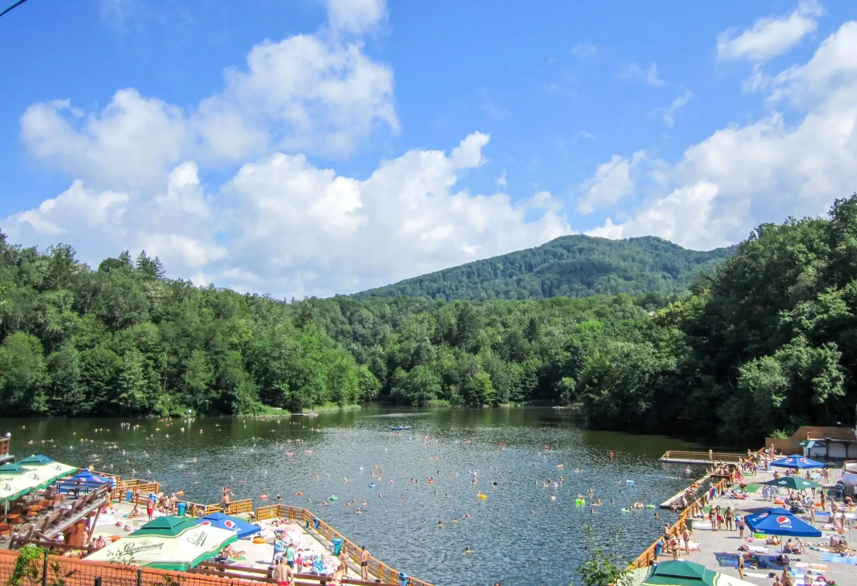 A picturesque lake enveloped by a charming promenade, where people swim and lounge leisurely, while lush trees grace the opposite shore and a verdant hill looms in the distance.