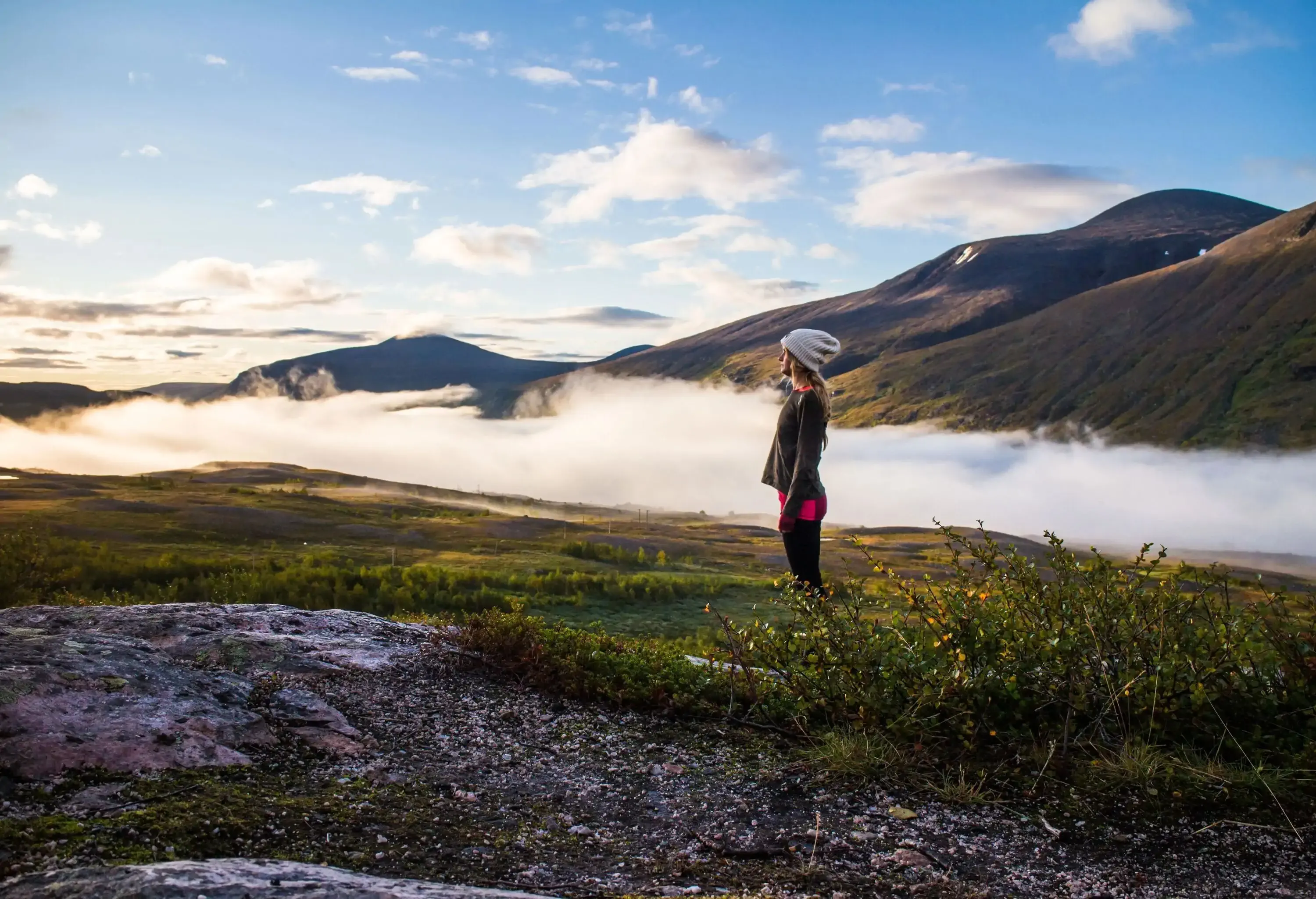 A woman stands on a lush landscape against the cloudy mountain range.
