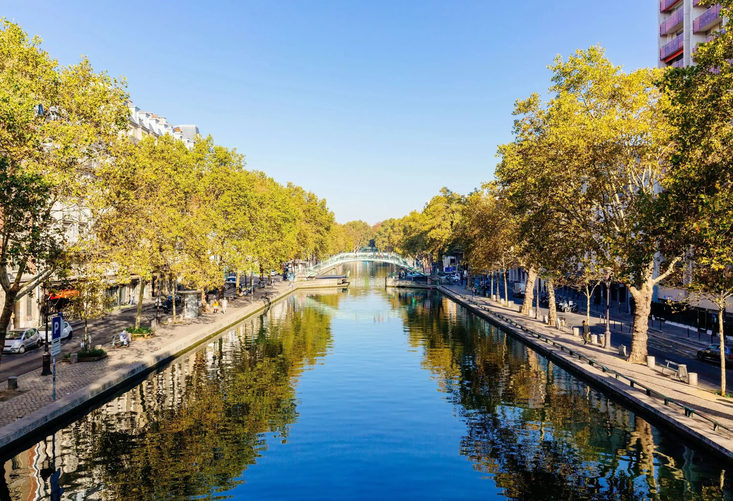 A calm water canal across a promenade lined with trees under a clear blue sky.