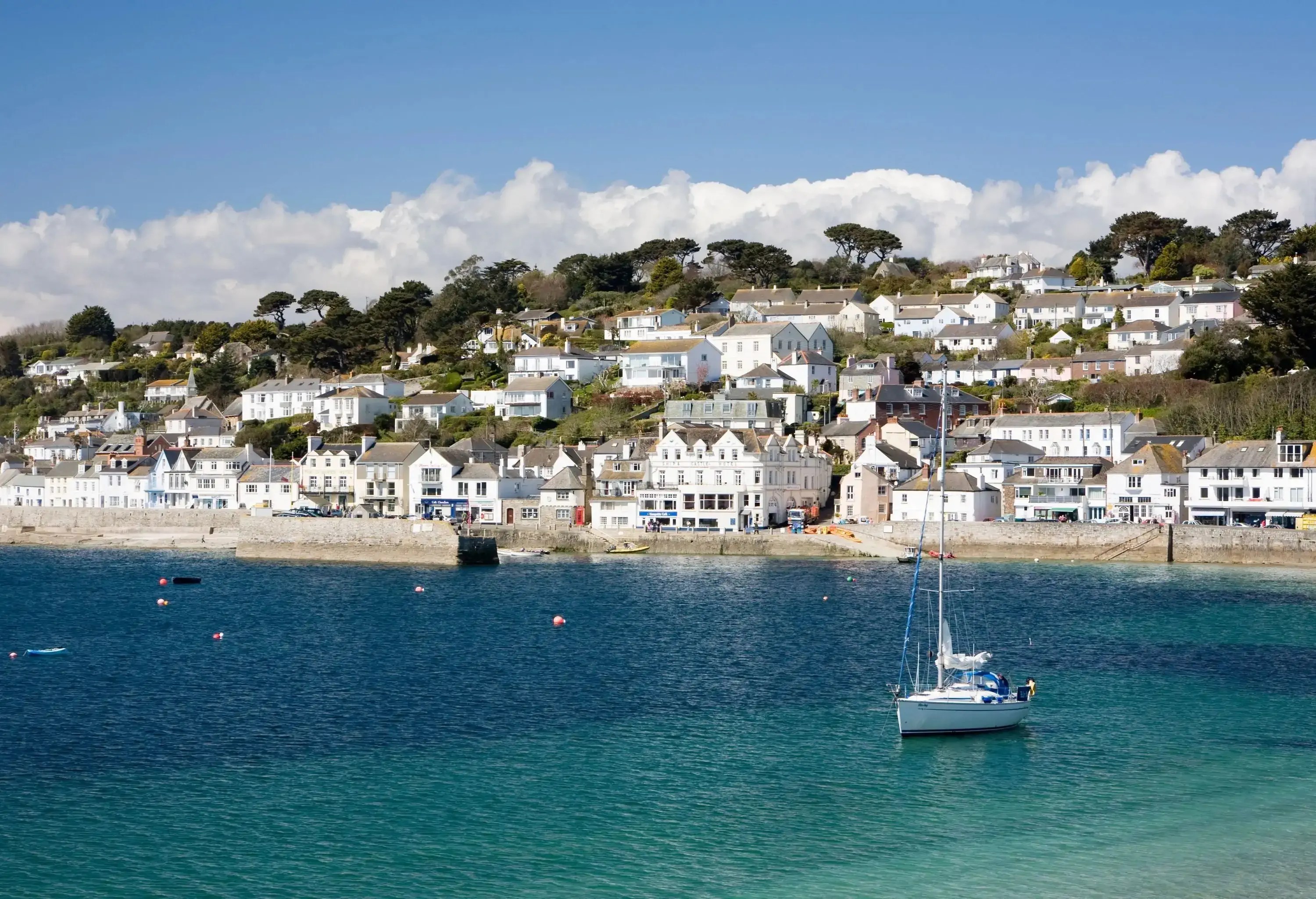 A small village of white houses nestled on the side of a hill with scattered trees along a clear, calm bay.