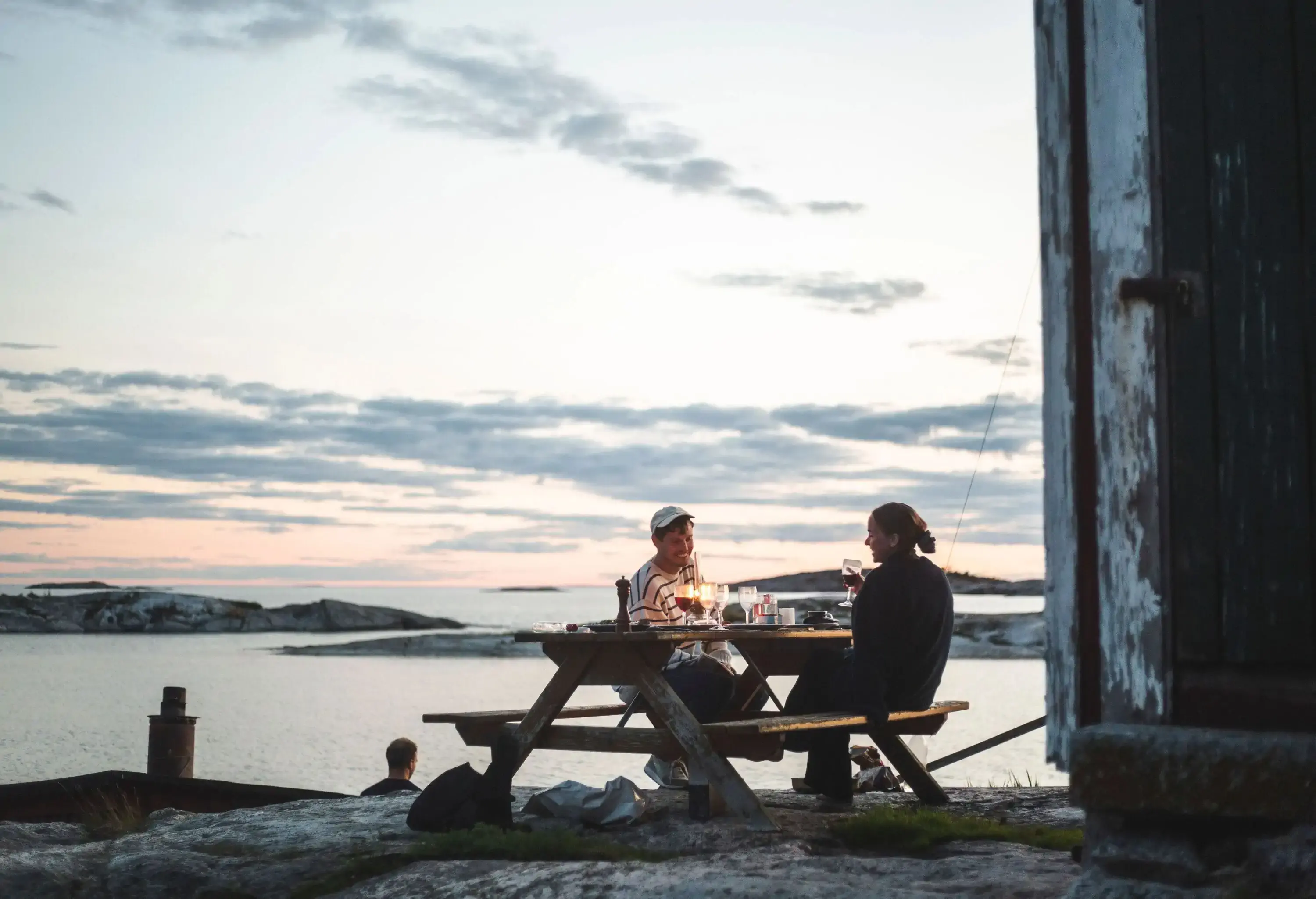 A couple sharing a drink sitting at a wooden table and chairs on a rocky outcrop overlooking the lake.