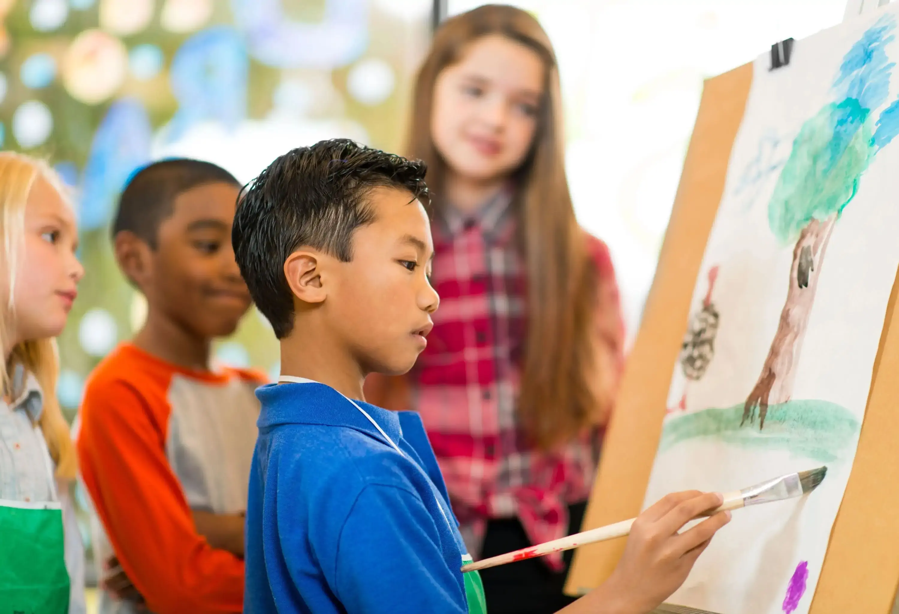 A boy in a blue shirt paints on a paper as his friends watch.