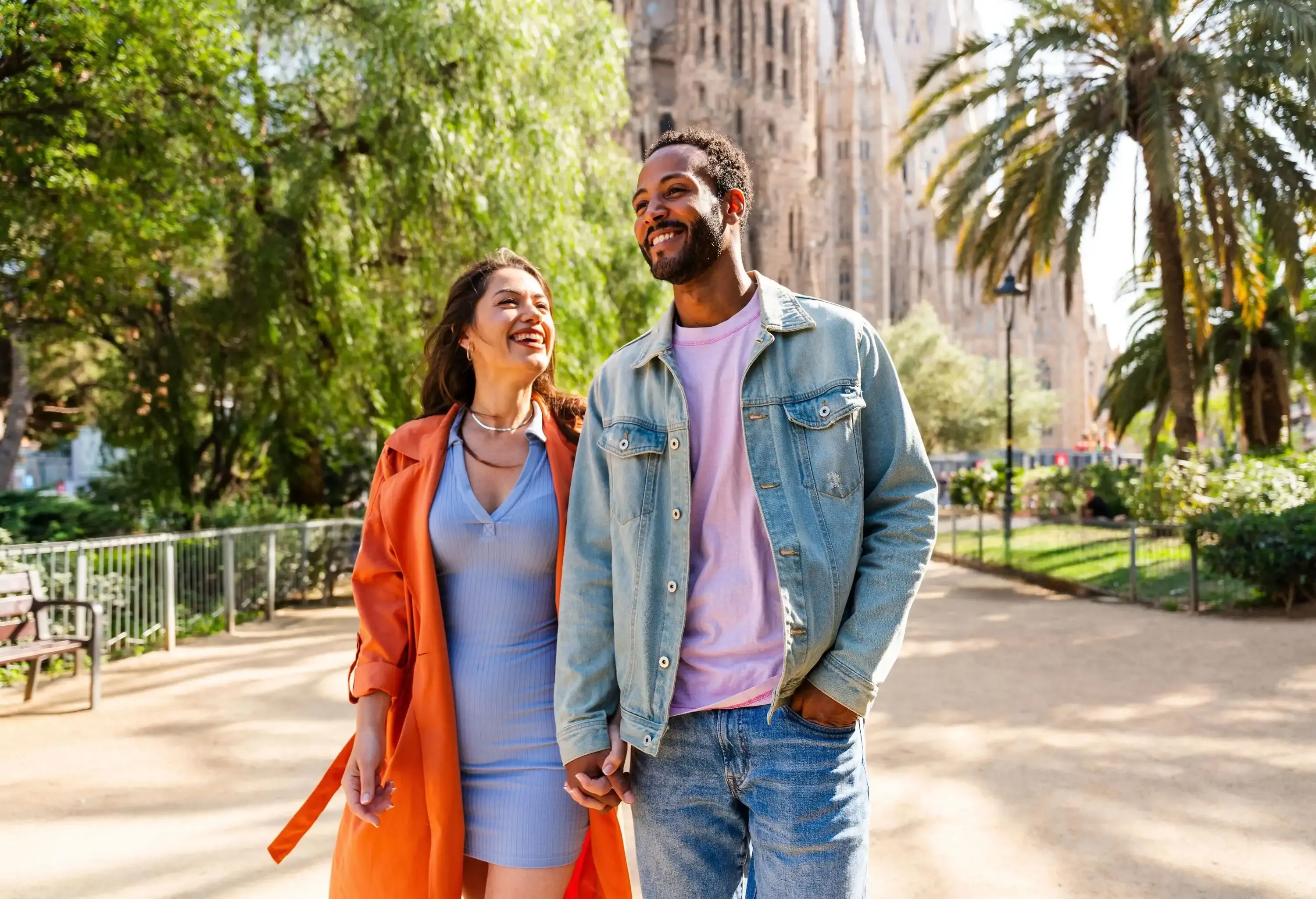 A happy couple strolling together on a path surrounded by greenery and a large, ornate building in the distance.
