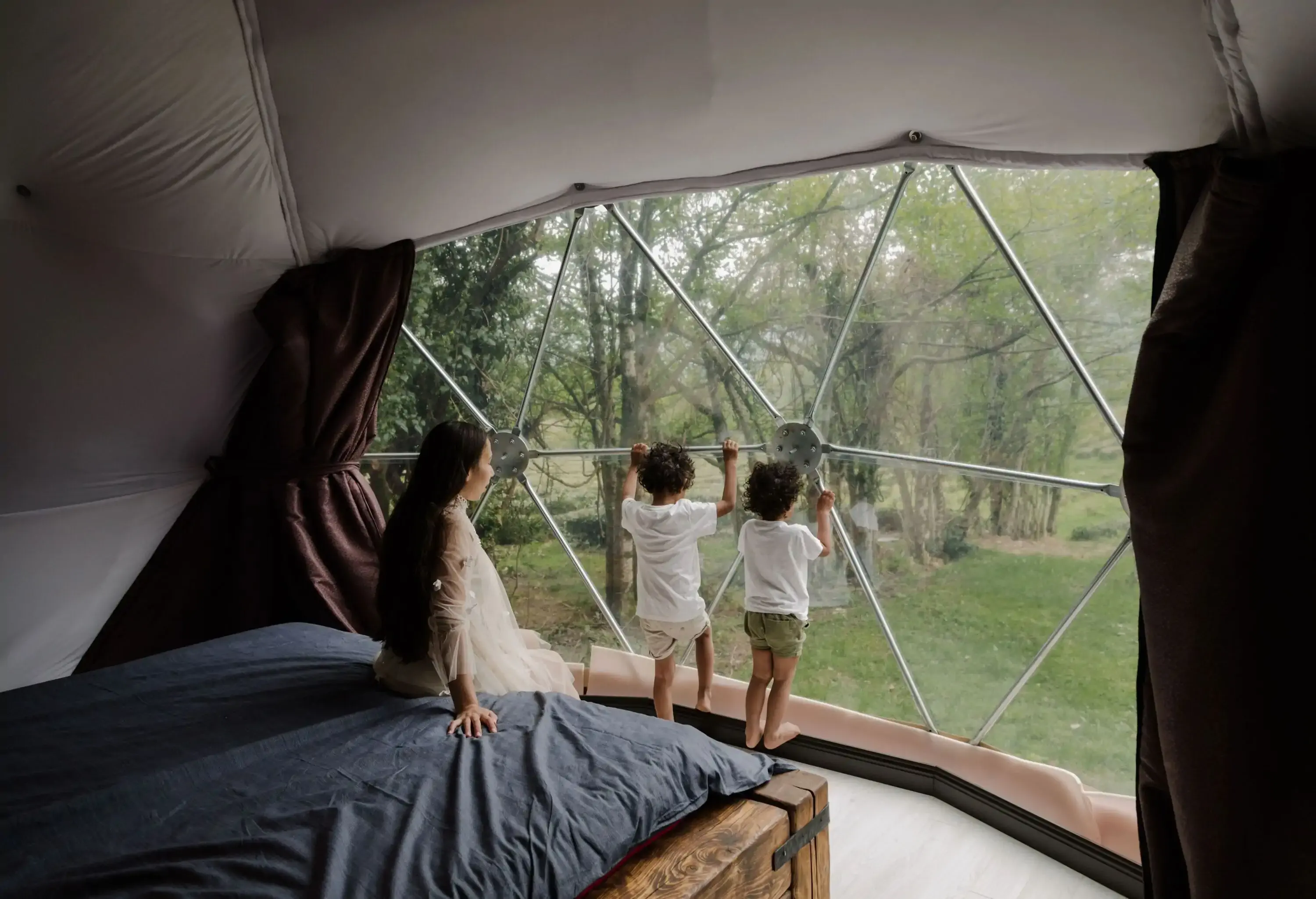 A mother is sitting on a bed next to two children gazing out a large window.