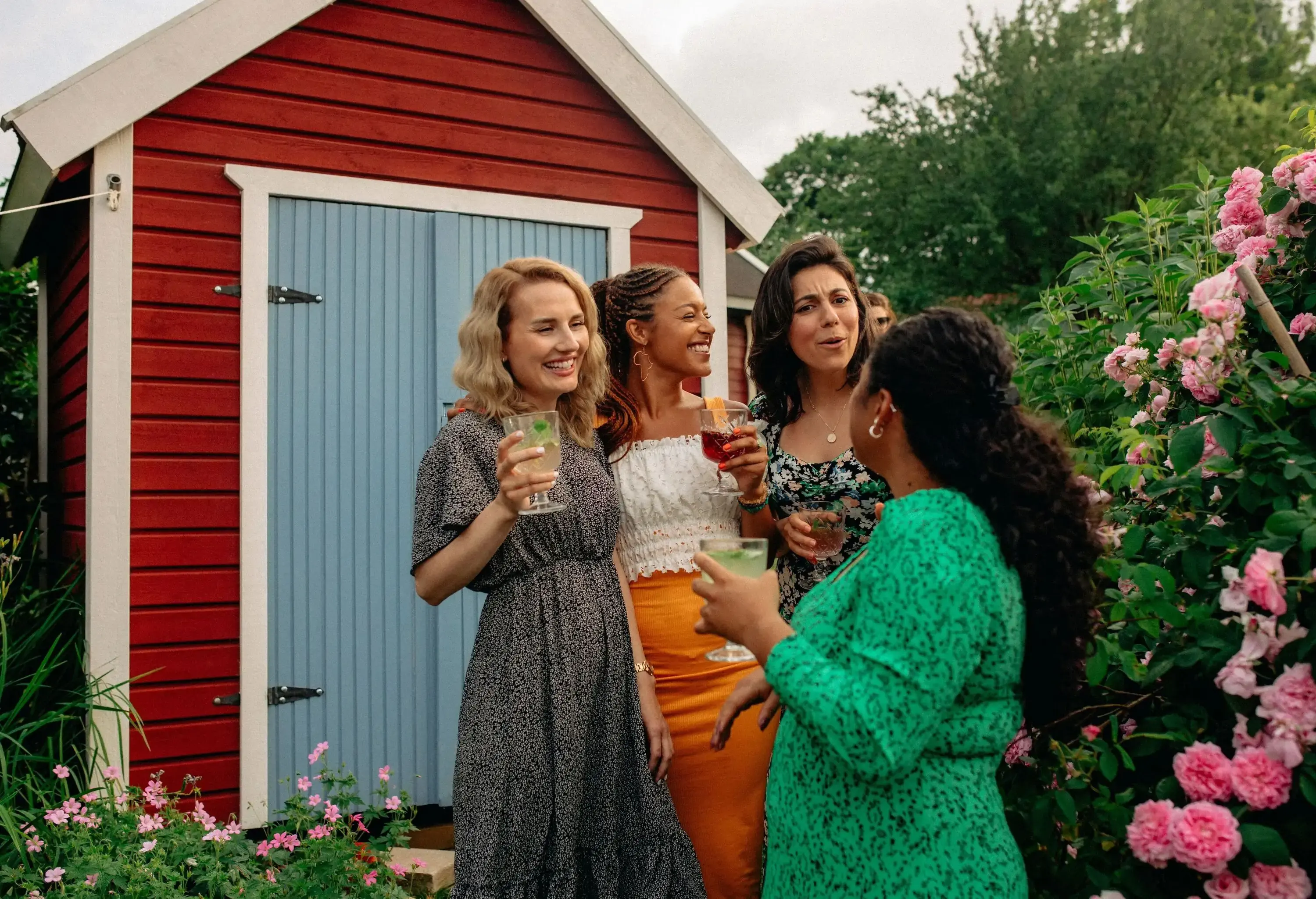 Four women laughing together while holding drinks, gathered outside a red garden shed, surrounded by pink flowers.