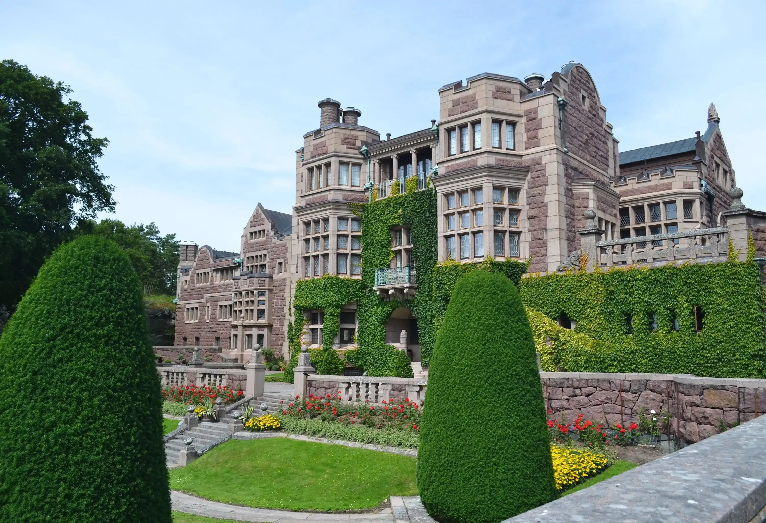 A Tudor-style castle with ornate chimneys and lush front gardens and shrubs.